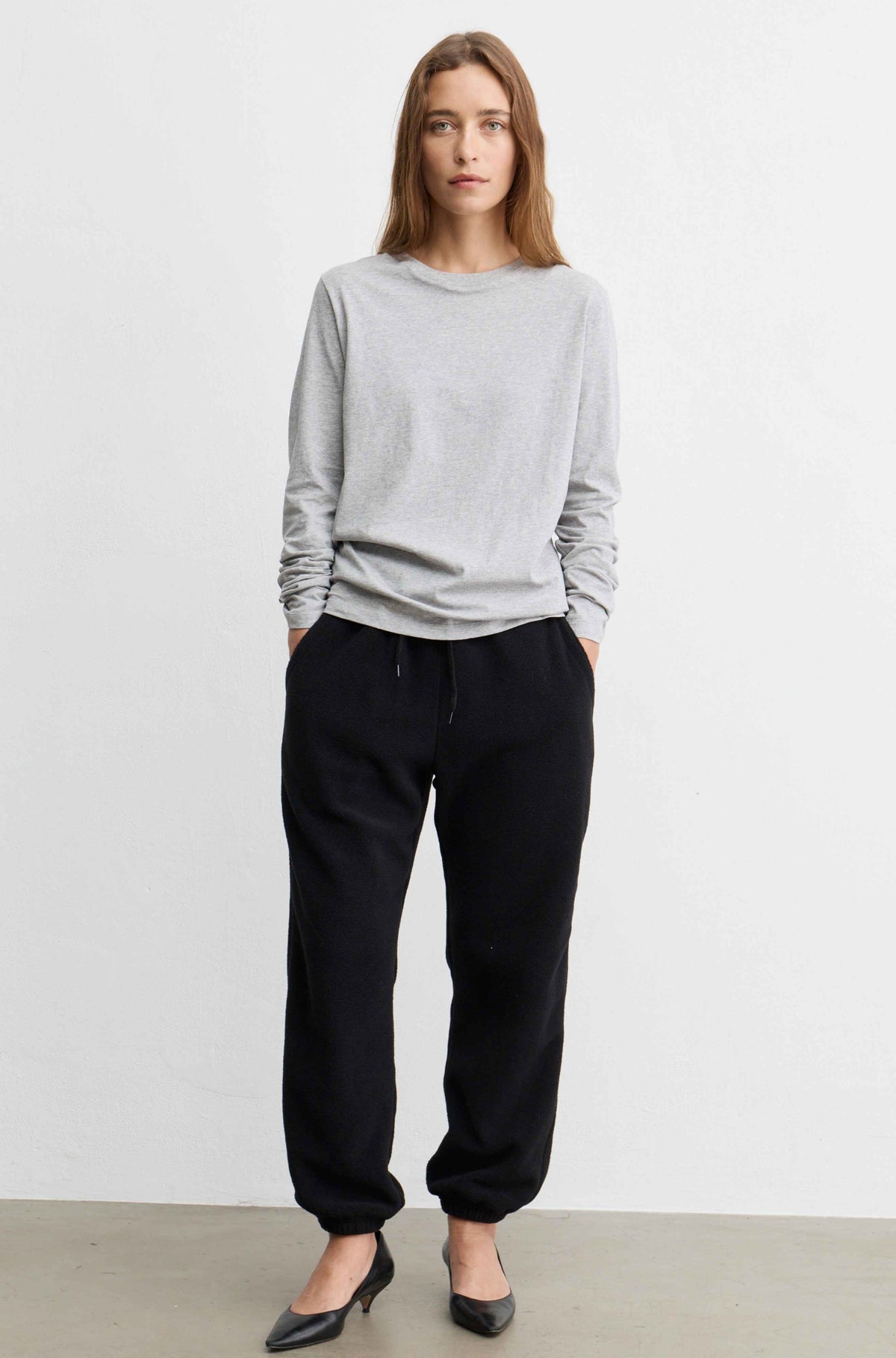A woman modeling a light gray long-sleeve crewneck sweatshirt and black jogger pants with black pointed-toe heels, standing against a plain white studio wall.