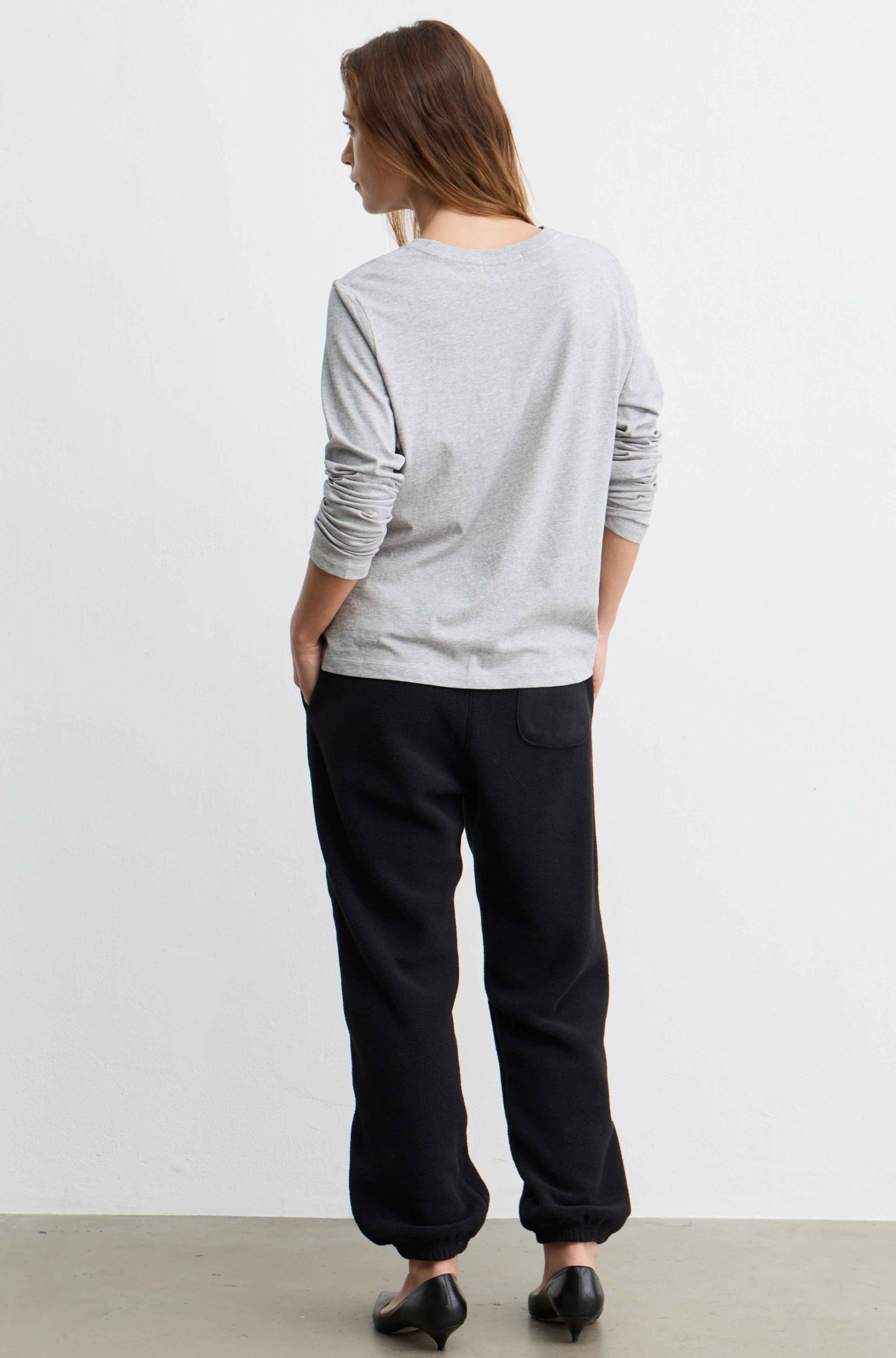 A woman is shown from behind wearing a light gray long-sleeve crewneck sweatshirt and black wide-leg sweatpants, standing against a plain white wall.
