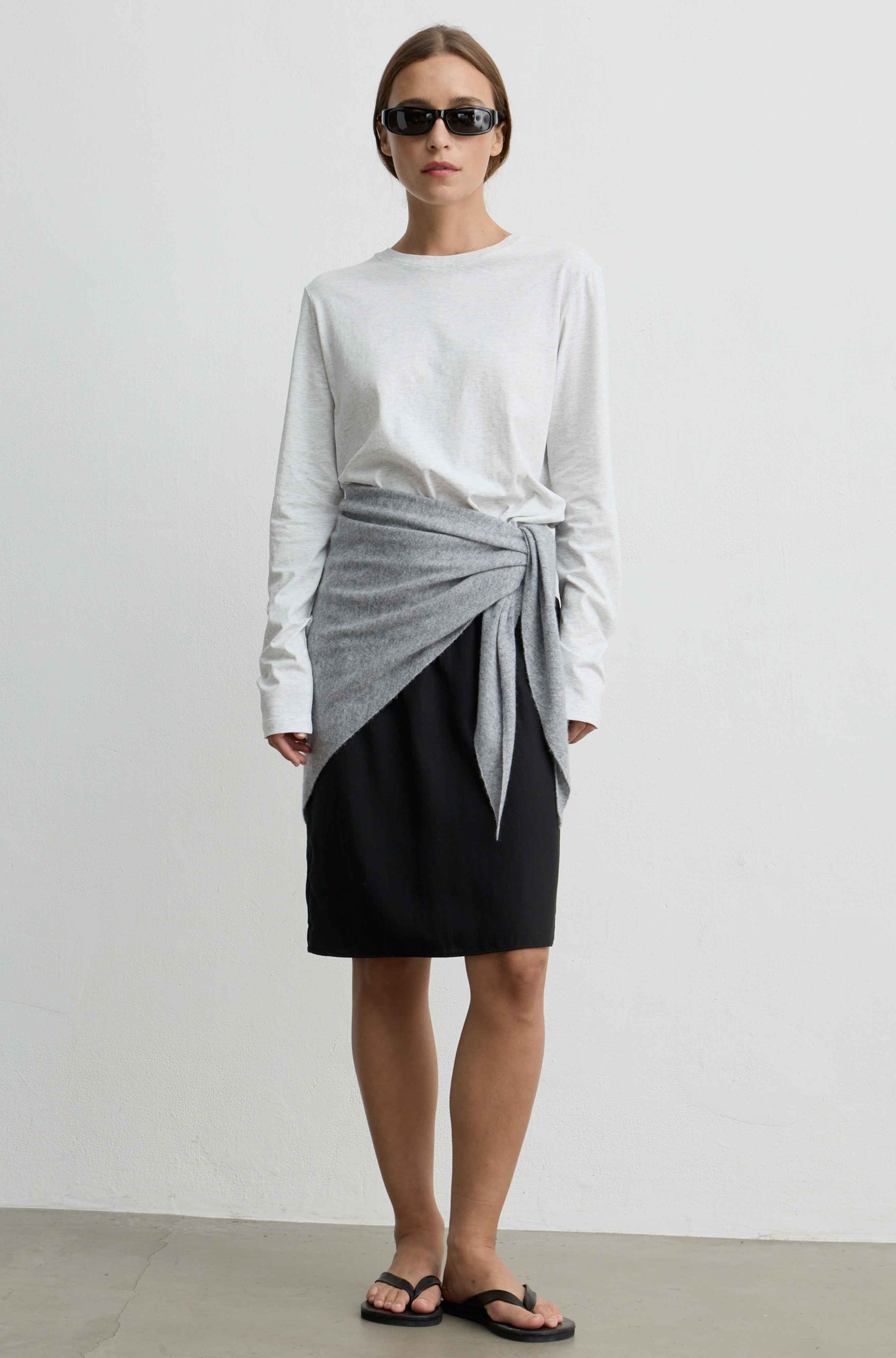 A model stands against a plain light gray wall wearing a white long-sleeve top, a black skirt, and a light gray wrap tied at the waist, with black sandals and sunglasses.