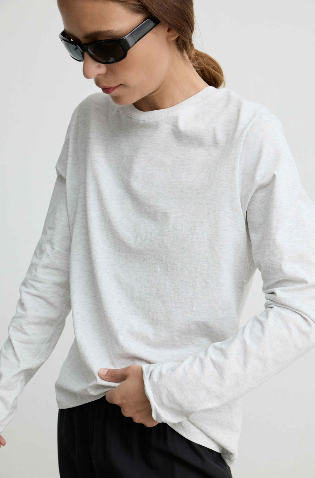 A woman wearing a light heather gray long-sleeve crewneck top and black sunglasses, shown from the chest down against a plain light background.