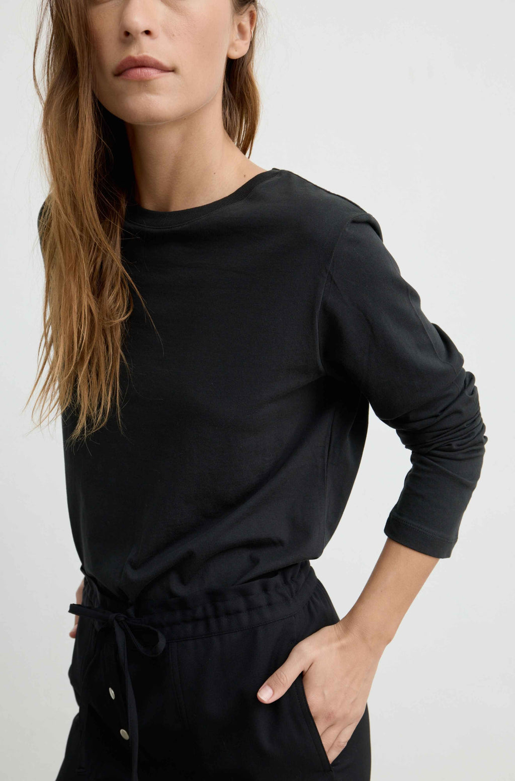 A woman wearing a black long-sleeve crewneck top and matching black drawstring pants, photographed against a plain light gray studio background.