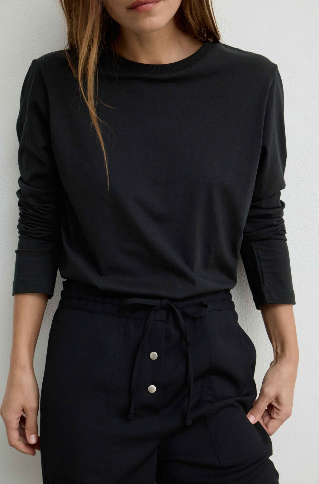 A woman wearing a black long-sleeve crewneck top and matching black drawstring pants, shown from the mouth down against a plain light background.