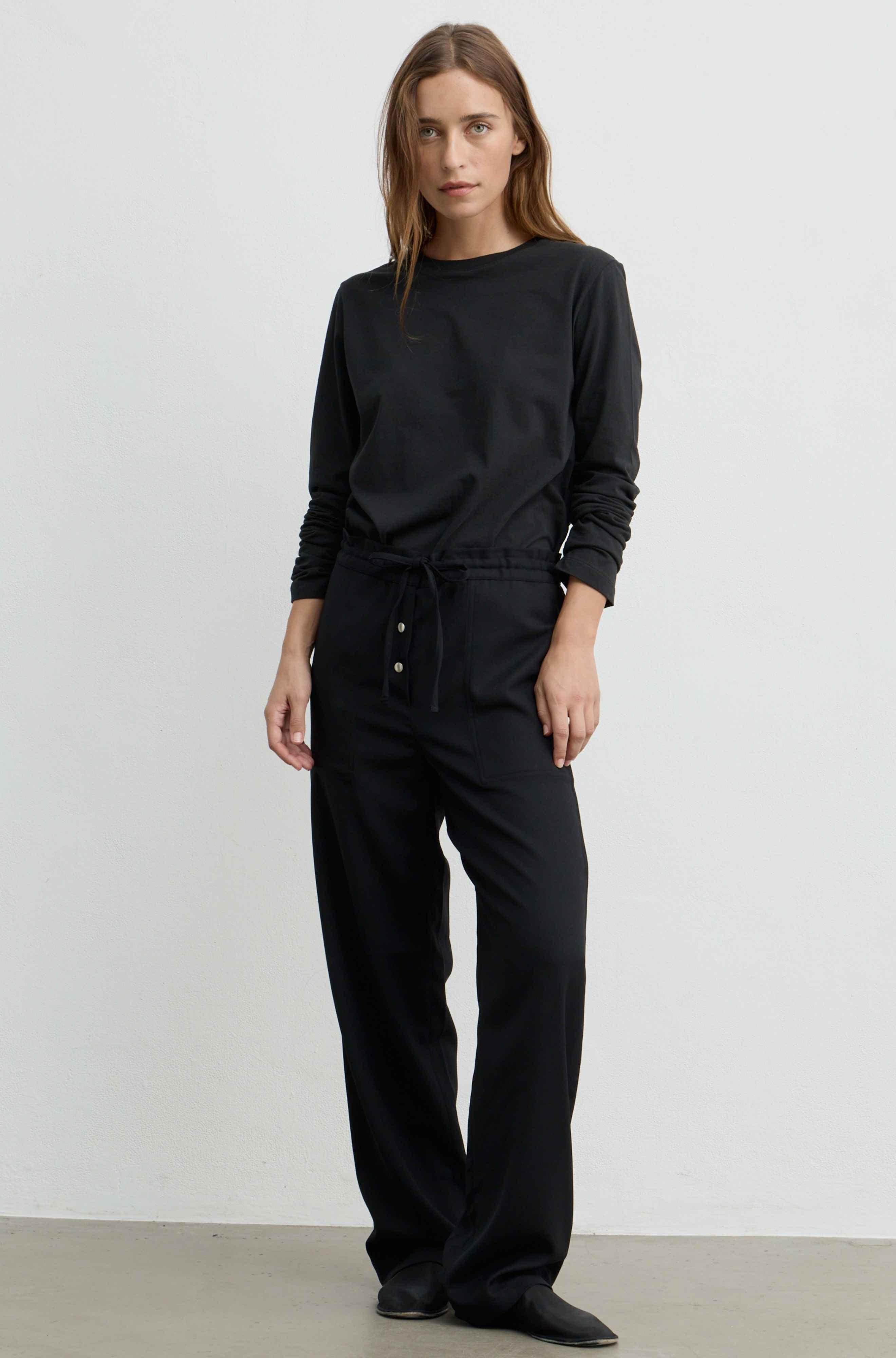 A woman modeling a black long-sleeve crewneck sweatshirt and matching black wide-leg drawstring pants with front buttons, standing against a plain light gray studio wall.