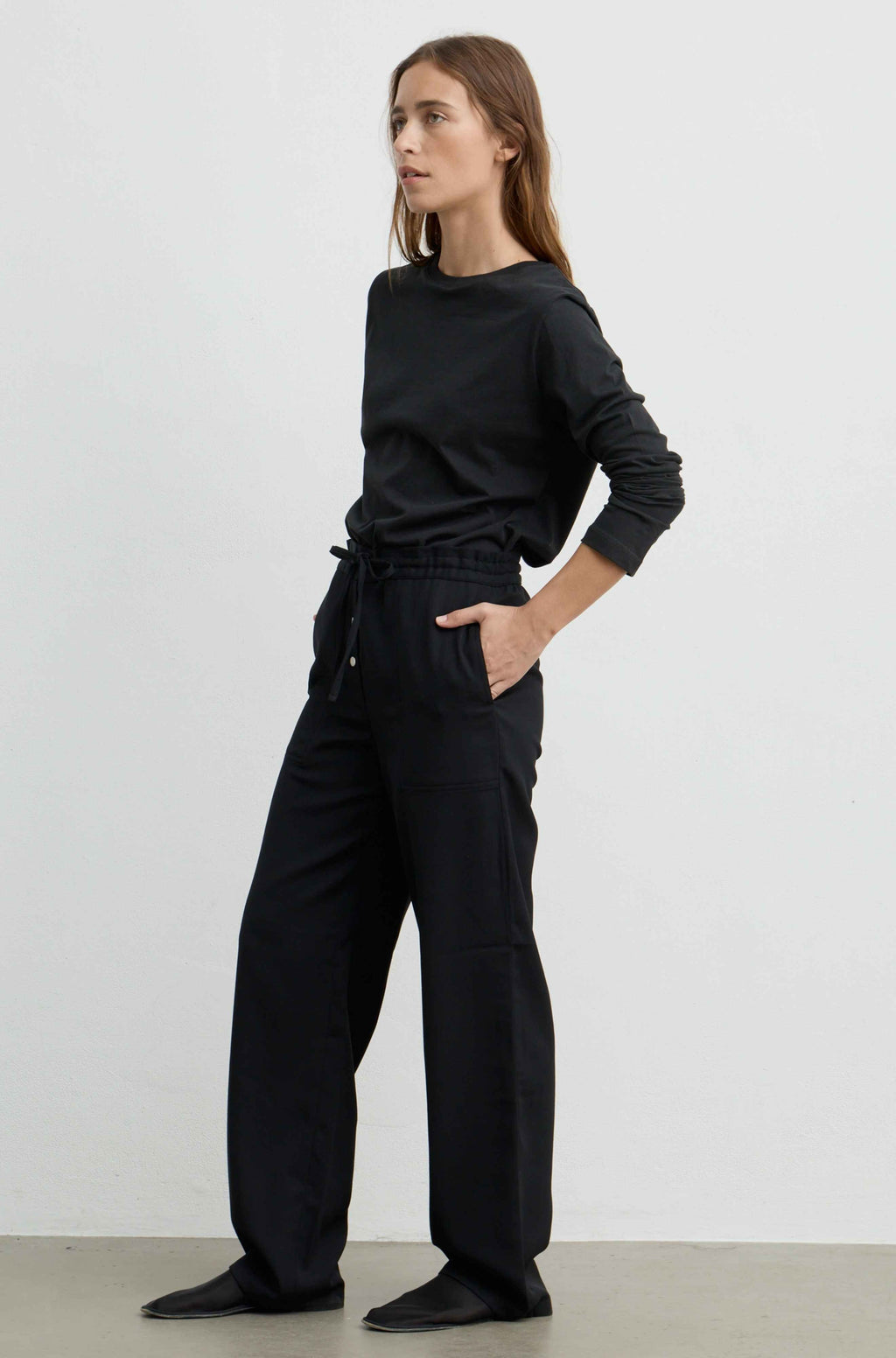A woman modeling a black long-sleeve top and black wide-leg drawstring pants against a plain light gray studio background.