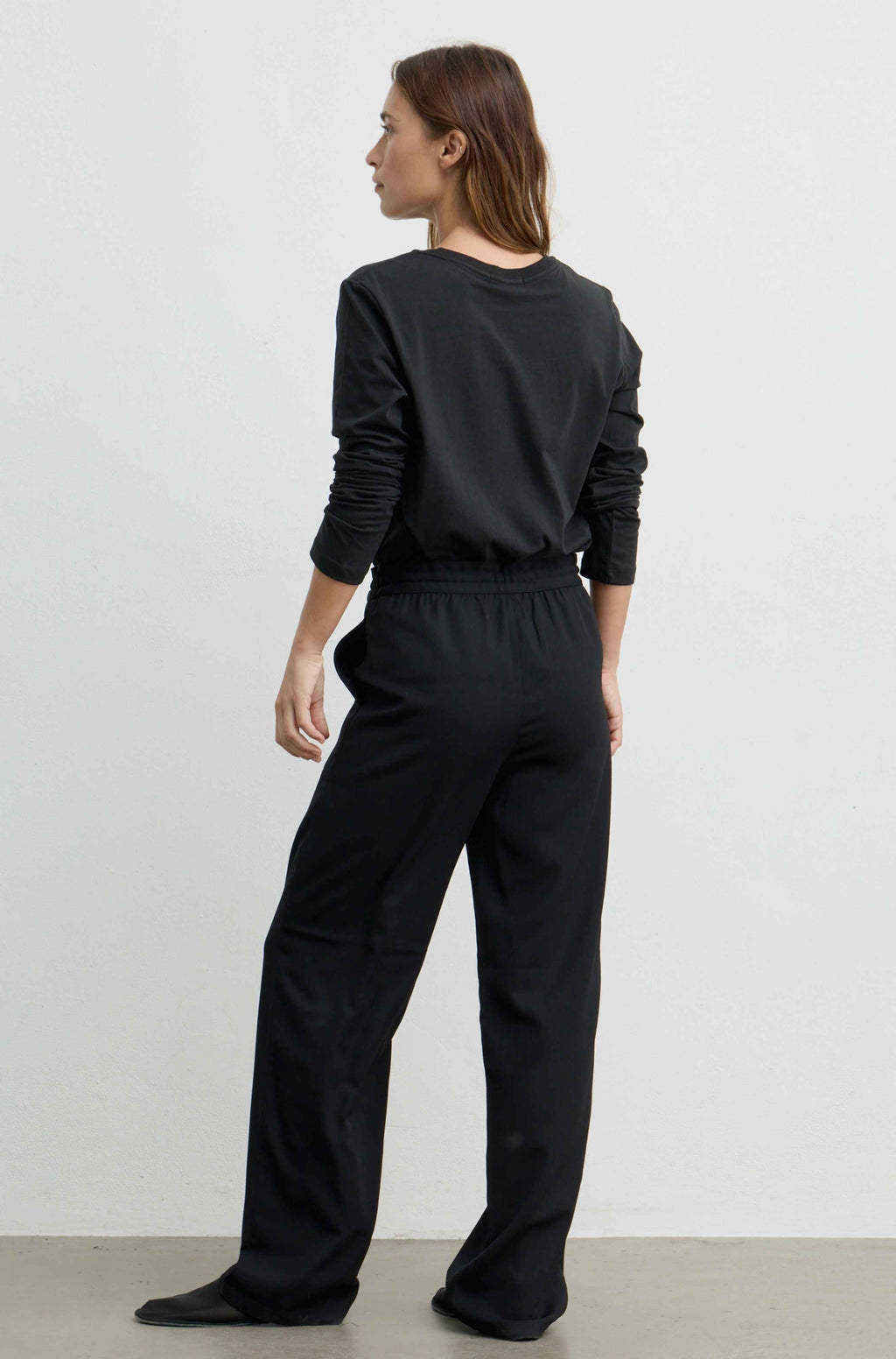 A woman wearing a black long-sleeve top and matching black wide-leg trousers, shown from the back against a plain light gray studio background.