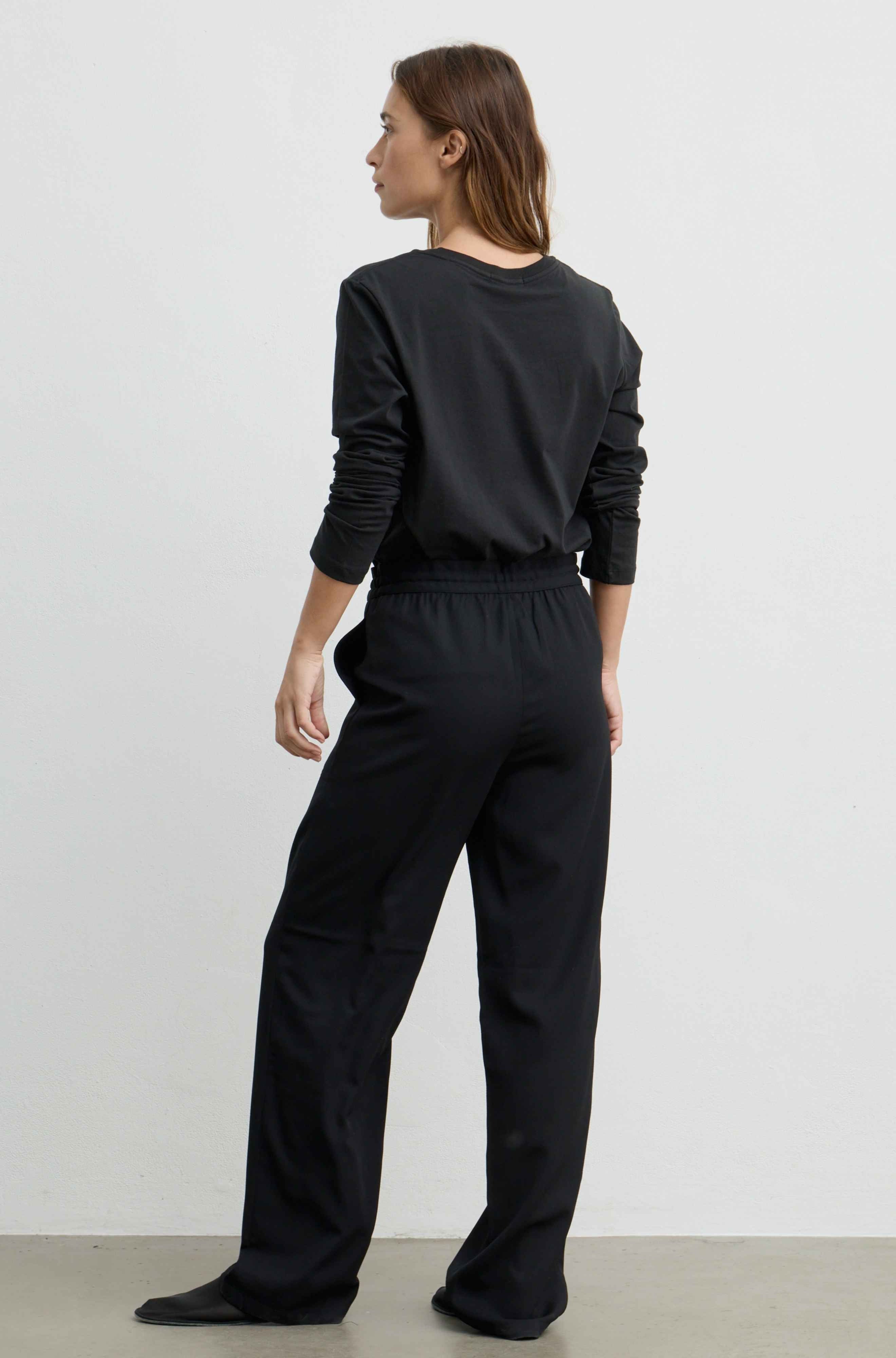 A woman wearing a black long-sleeve top and matching black wide-leg trousers, shown from the back against a plain light gray studio background.