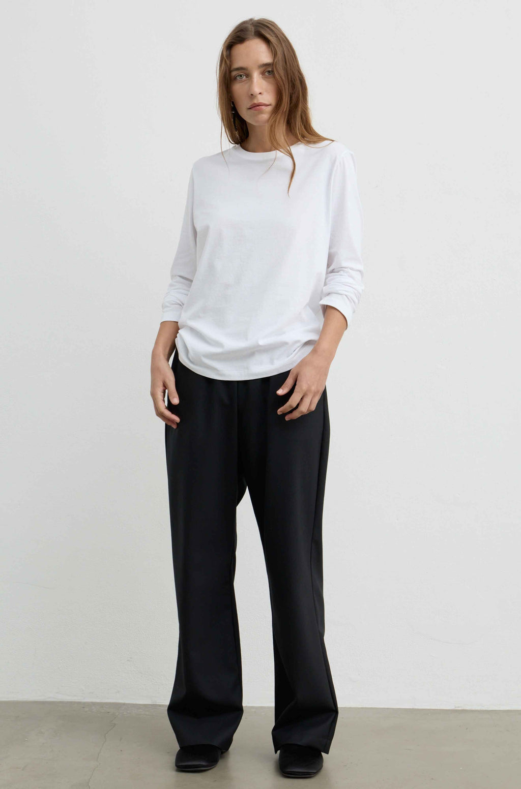 A woman modeling a white long-sleeve crewneck top and black wide-leg trousers against a plain off-white studio background.