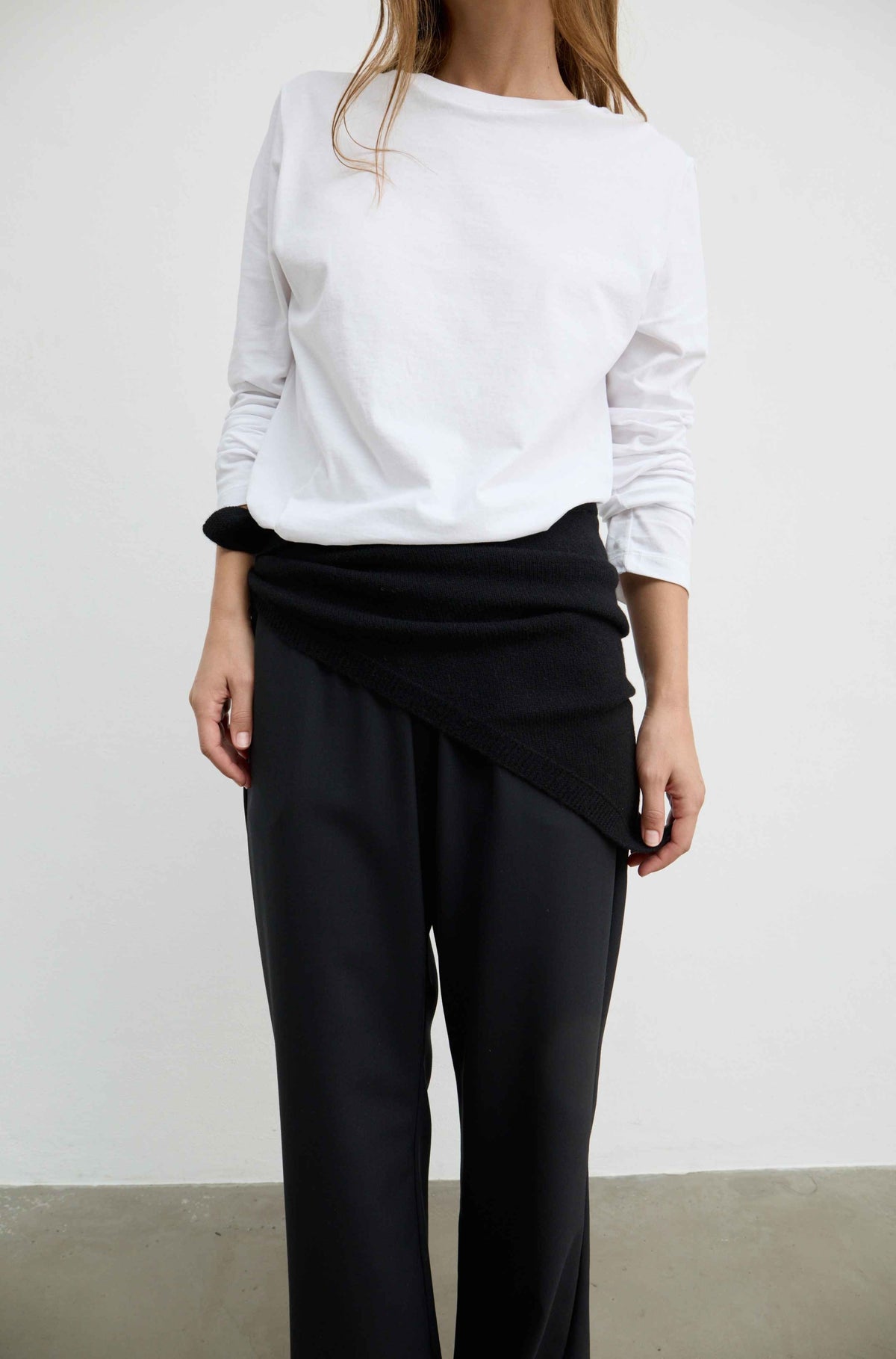A model wearing a white long-sleeve top and black wrap-style trousers, standing against a plain light gray wall.