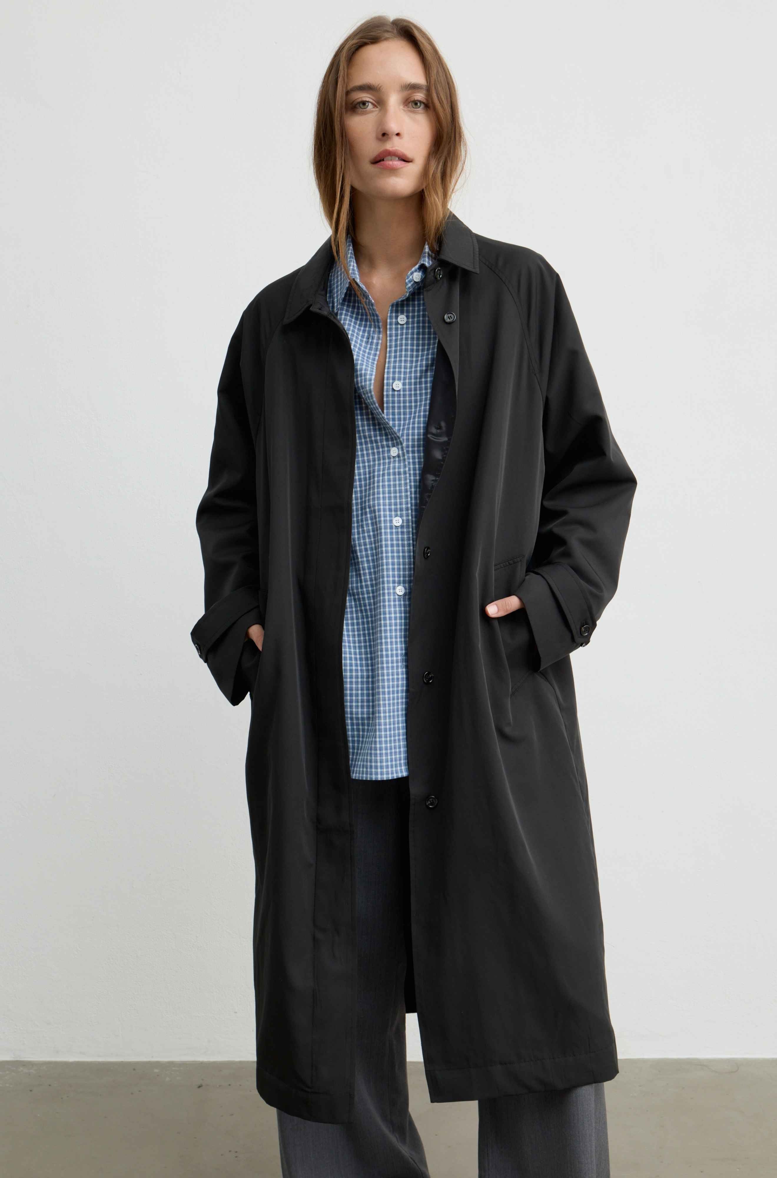 A woman stands against a plain light gray wall wearing a long black coat over a blue-and-white gingham button-up shirt and dark gray pants.