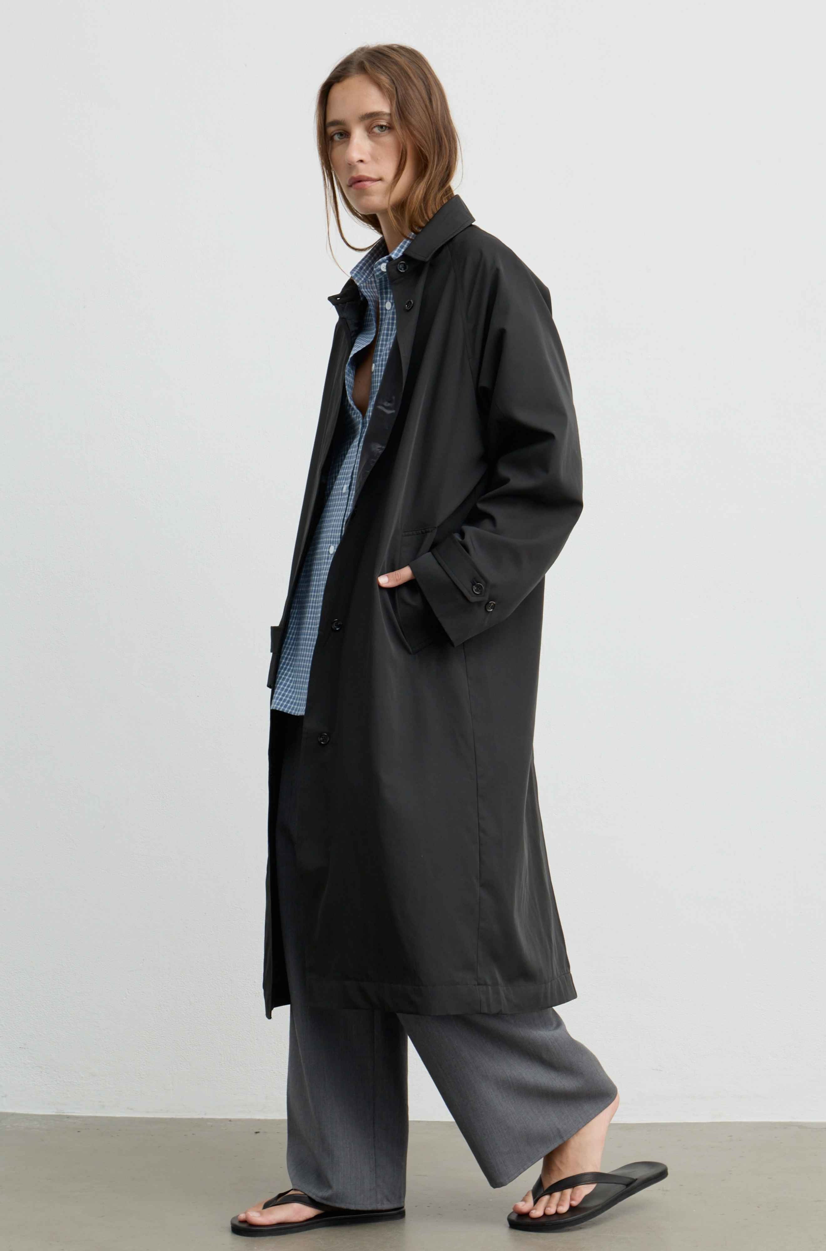A model wearing a long black trench coat over a blue and white checkered shirt, gray wide-leg pants, and black flip-flops, standing against a plain light gray wall.