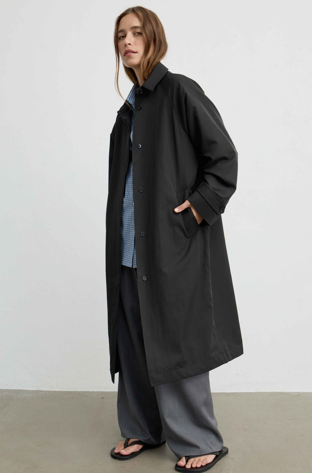 A woman modeling a long black coat over a blue striped shirt and gray wide-leg pants, standing against a plain light gray wall.