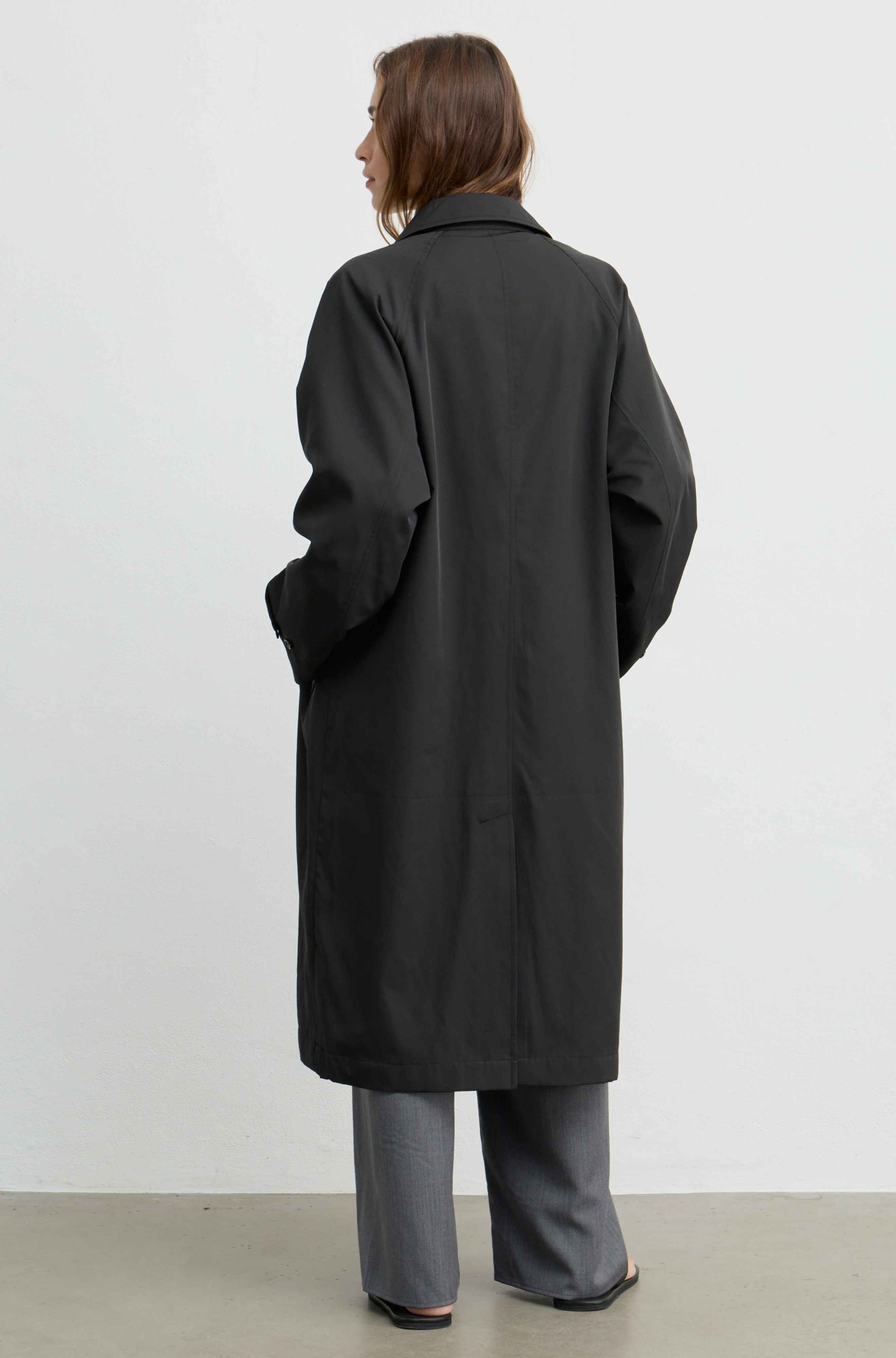 A woman viewed from the back wearing a long black coat over gray wide-leg pants, standing against a plain light gray studio wall.