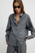 A woman wearing a loose-fitting, long-sleeve gray button-up shirt and matching gray trousers, styled with black sunglasses against a plain light gray studio background.