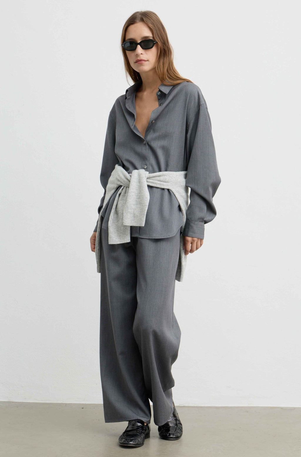 A model wearing a gray two-piece outfit consisting of a long-sleeve button-up shirt and matching wide-leg trousers, styled with a light gray tie-waist sash and black loafers.