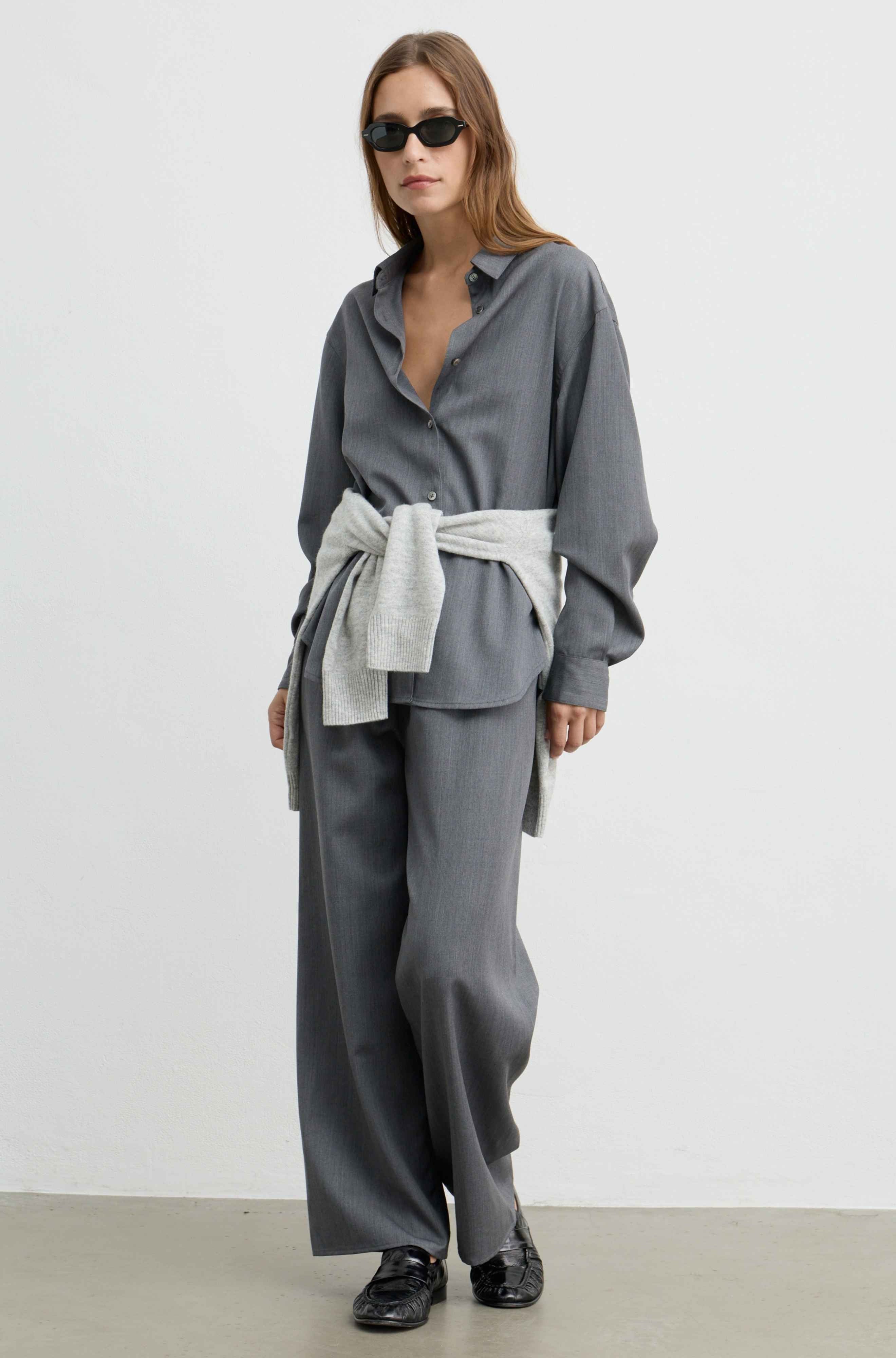 A model wearing a gray two-piece outfit consisting of a long-sleeve button-up shirt and matching wide-leg trousers, styled with a light gray tie-waist sash and black loafers.