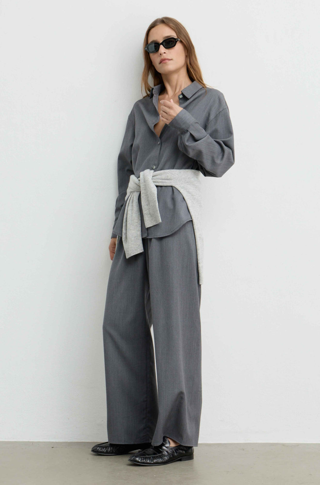 A woman modeling a gray long-sleeve collared shirt and matching wide-leg trousers, styled with a light gray tied sash at the waist and black sunglasses, standing against a plain light gray wall.