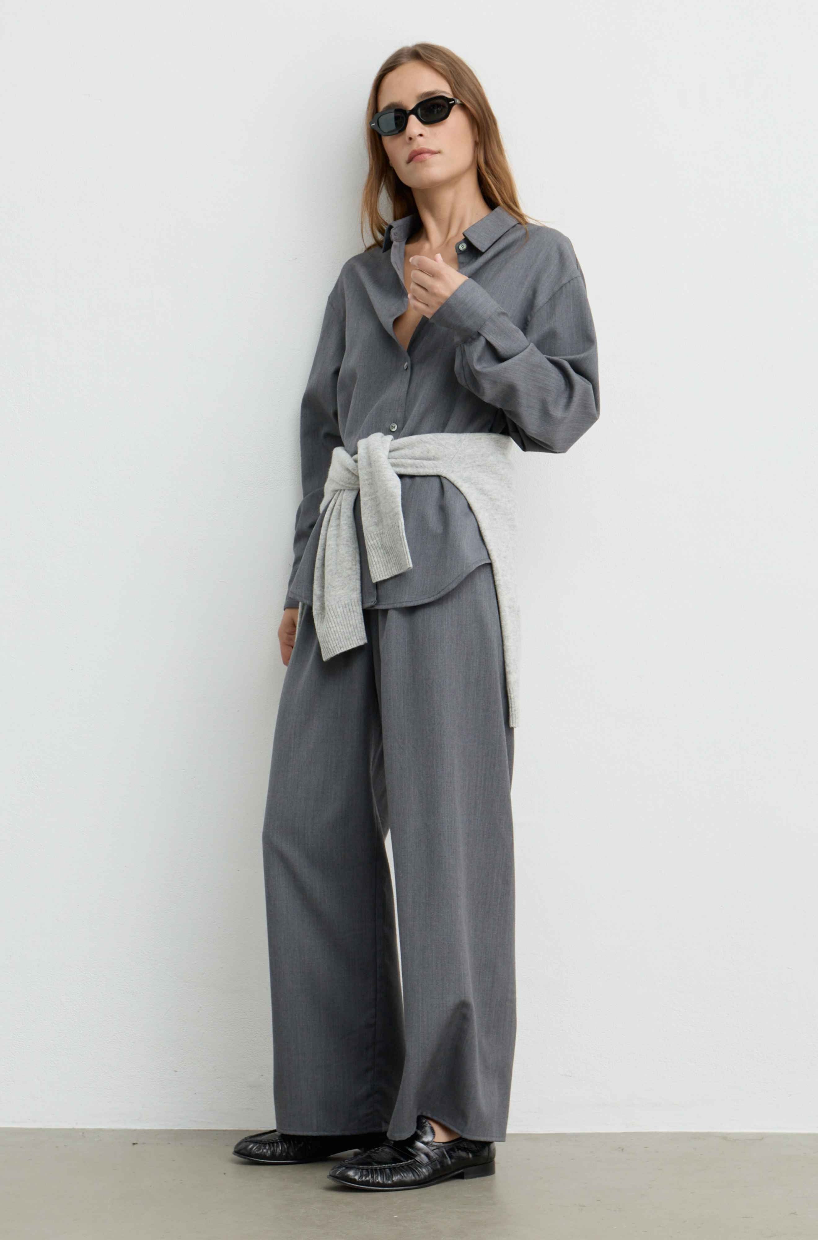 A woman modeling a gray long-sleeve collared shirt and matching wide-leg trousers, styled with a light gray tied sash at the waist and black sunglasses, standing against a plain light gray wall.