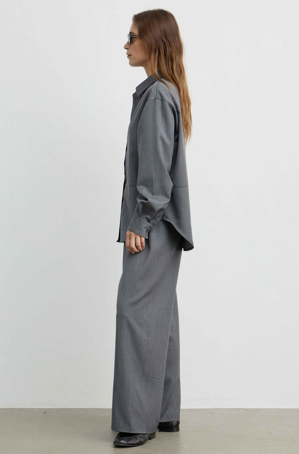 A woman stands in profile wearing a loose-fitting gray long-sleeve button-up shirt and matching wide-leg gray trousers, photographed against a plain light gray studio wall.