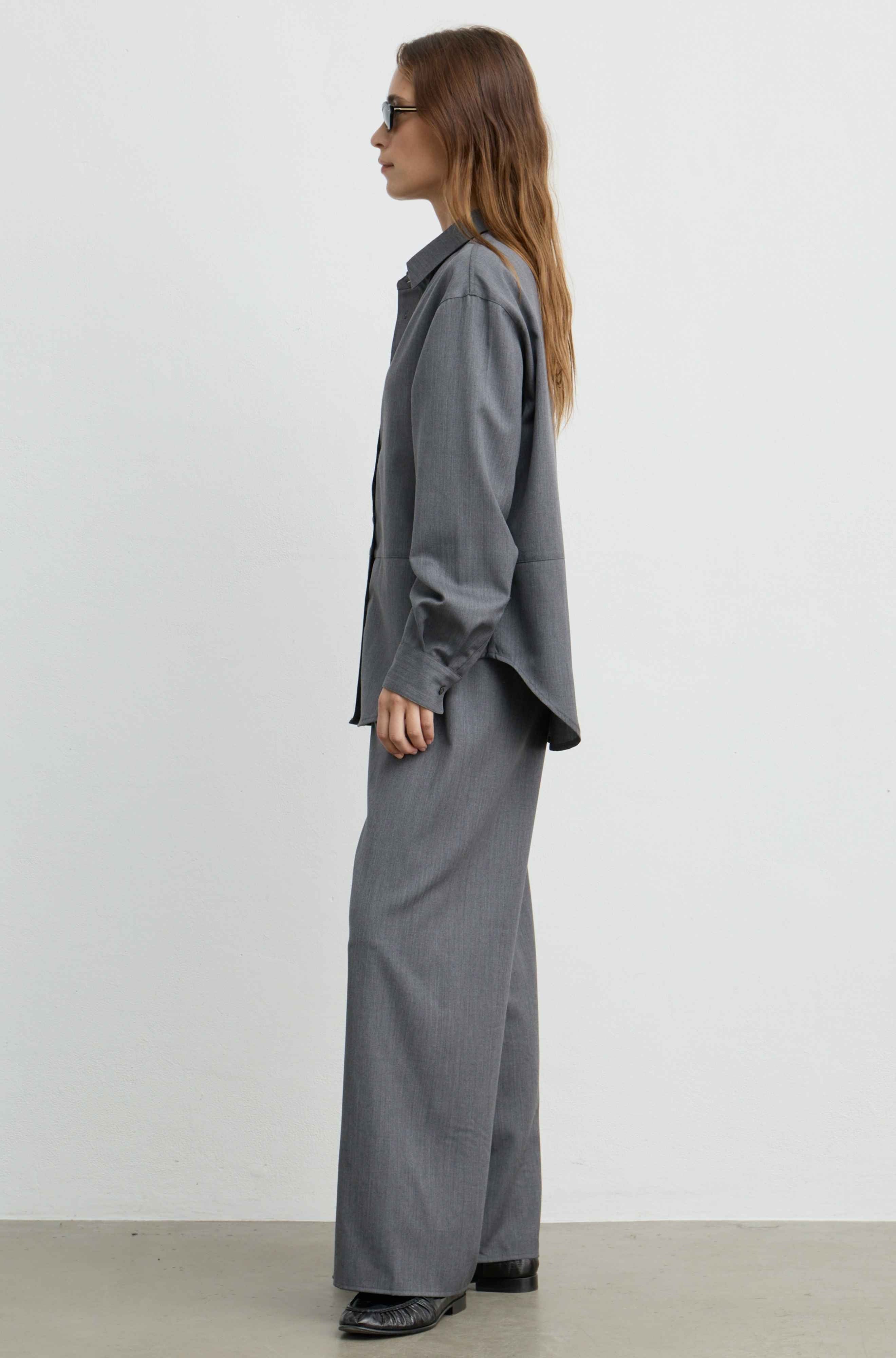A woman stands in profile wearing a loose-fitting gray long-sleeve button-up shirt and matching wide-leg gray trousers, photographed against a plain light gray studio wall.