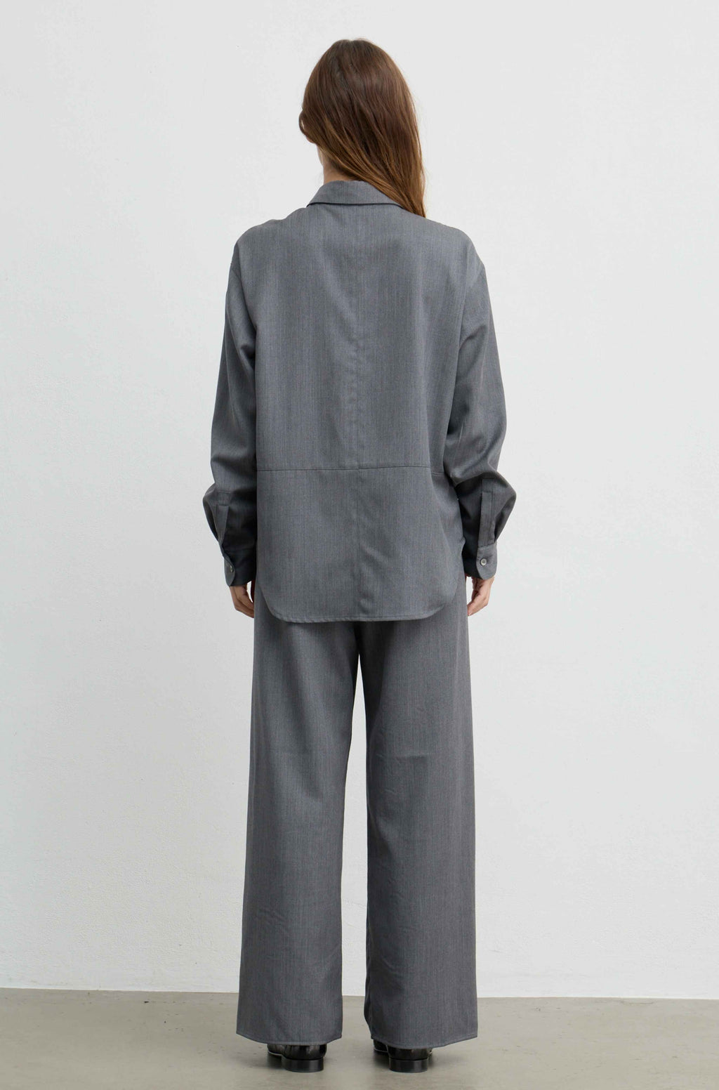 A model viewed from the back wearing a matching gray two-piece suit consisting of a long-sleeve button-down shirt and wide-leg trousers, set against a plain white studio background.