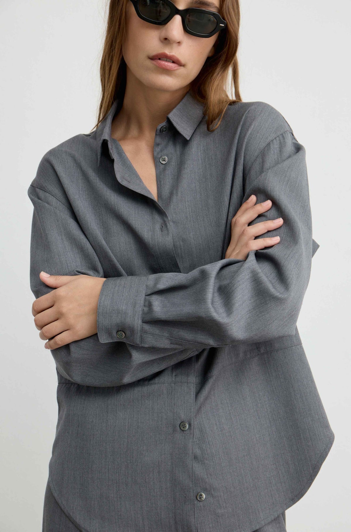 A woman wearing an oversized dark gray button-down shirt with long sleeves and a relaxed fit, styled with black sunglasses against a plain light gray background.