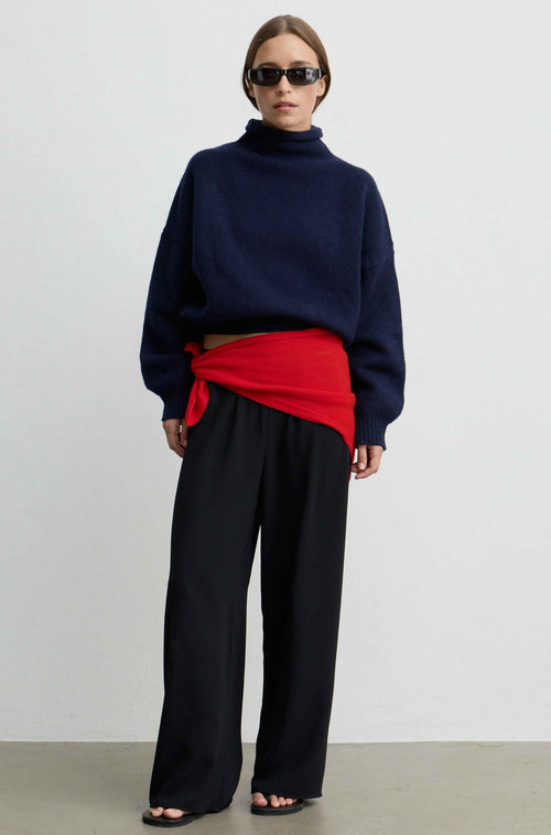 Woman wearing a navy knit sweater with a mock turtleneck and a red scarf tied around her waist with black pants against a light background