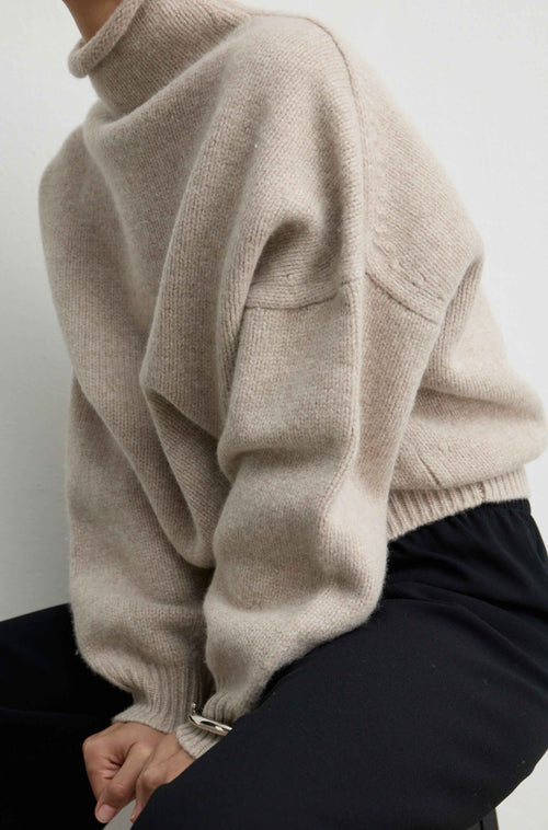 Close up of a woman wearing a beige knit wool sweater and black pants on a light grey background
