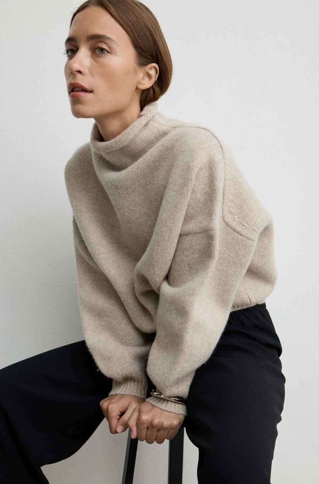 Woman sitting on a chair wearing a beige knit sweater with turtleneck and black pants sitting on a chair