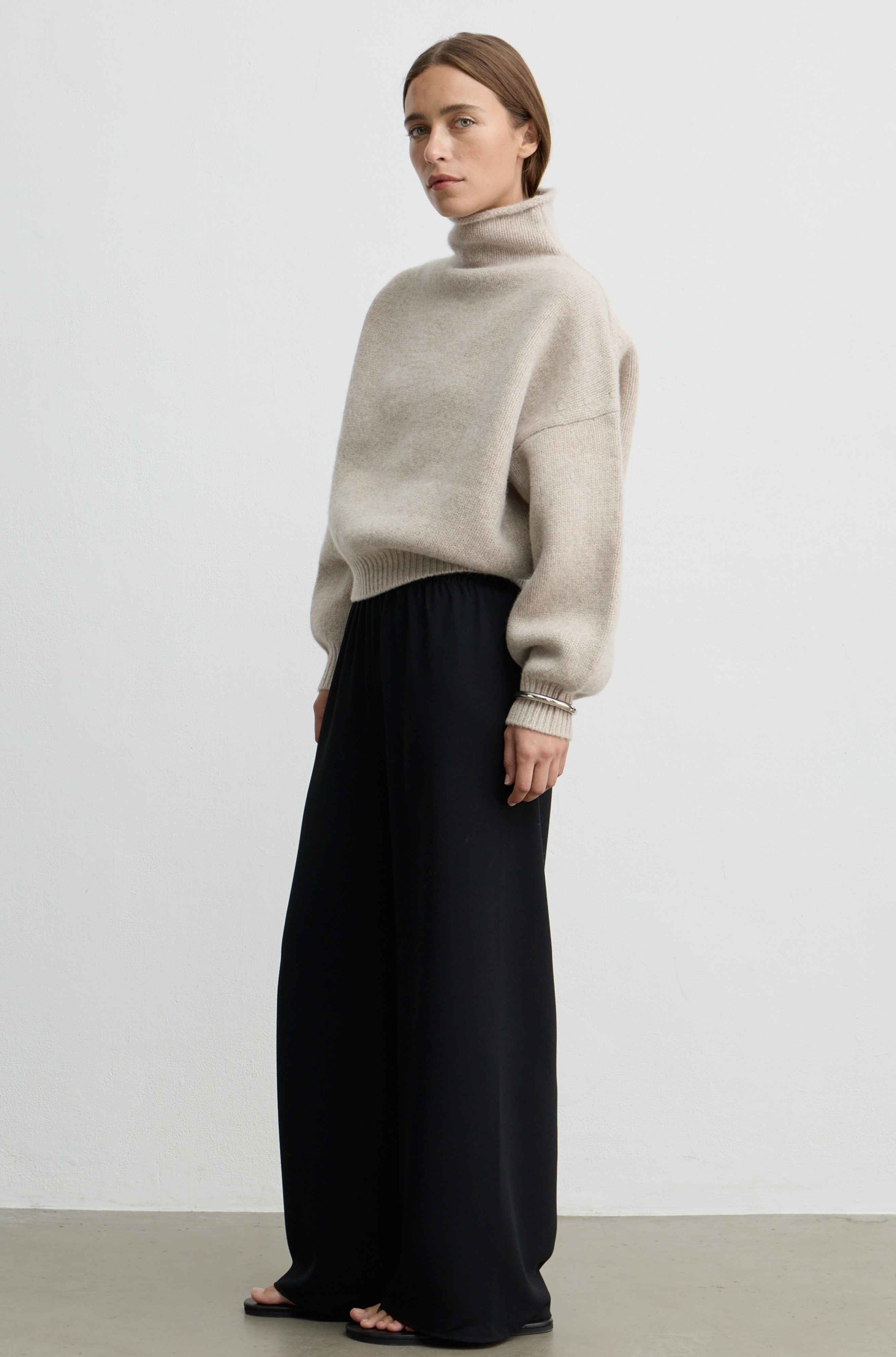 Woman wearing a beige oatmeal wool sweater with black pants against a light grey background