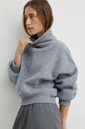 A woman wearing a light gray oversized knit sweater with a high folded collar and ribbed cuffs, paired with grey pants, photographed against a plain light gray wall.