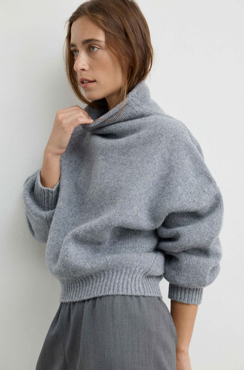 A woman wearing a light gray oversized knit sweater with a high folded collar and ribbed cuffs, paired with grey pants, photographed against a plain light gray wall.