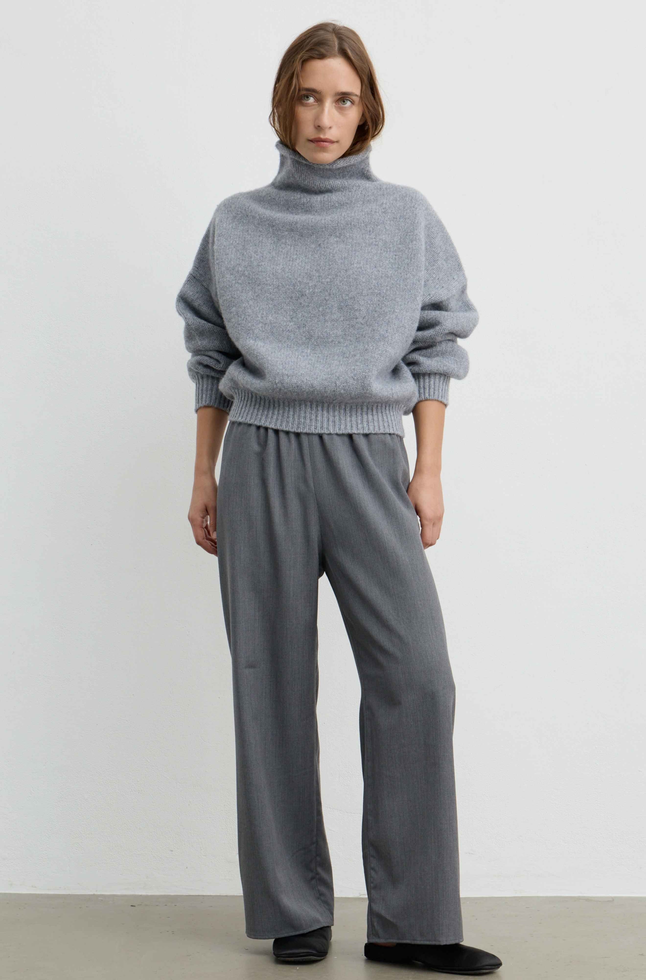 A woman wearing a light gray oversized knit sweater with a high folded collar and ribbed cuffs, paired with grey pants, photographed against a plain light gray wall.