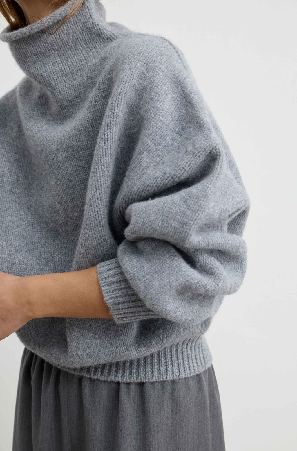 A woman wearing a light gray oversized knit sweater with a high folded collar and ribbed cuffs, paired with grey pants, photographed against a plain light gray wall.