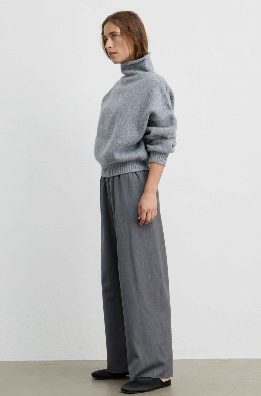 A woman wearing a light gray oversized knit sweater with a high folded collar and ribbed cuffs, paired with a matching gray skirt, photographed against a plain light gray wall.