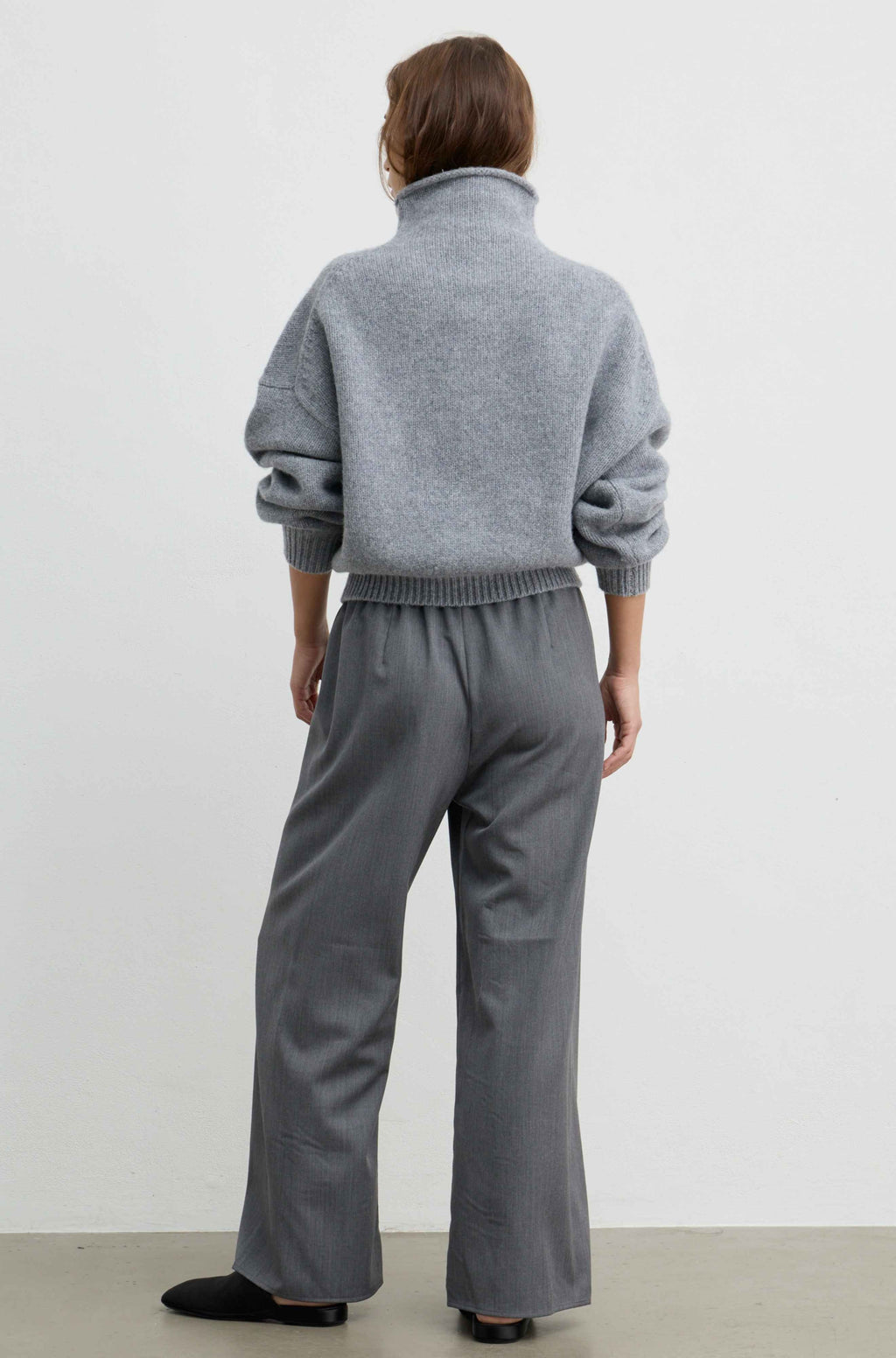 A woman wearing a light gray oversized knit sweater with a high folded collar and ribbed cuffs, paired with a matching gray skirt, photographed against a plain light gray wall.