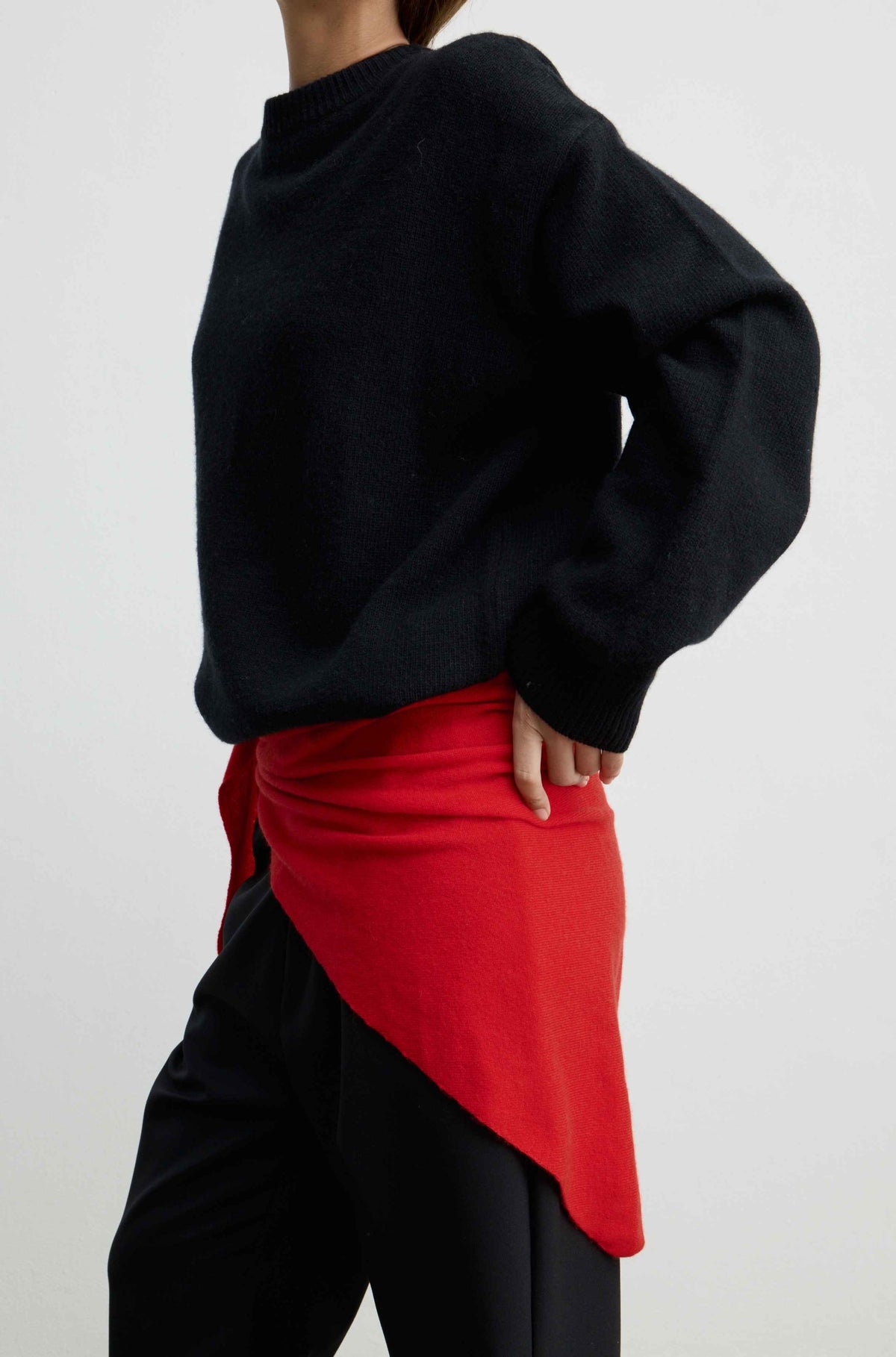 A person shown from the back wearing an oversized black knit sweater, black pants, and a bright red asymmetrical wrap or skirt detail tied at the waist, against a plain light gray studio background.