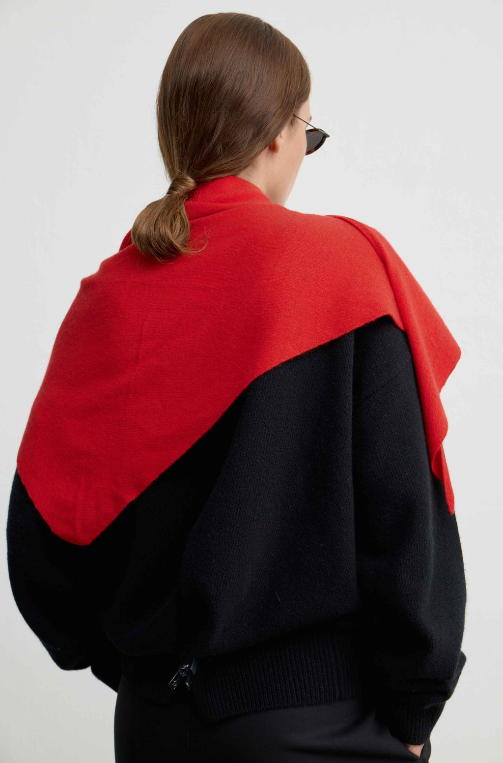 A woman shown from behind wearing a large red wrap or shawl draped over a black sweater against a plain light gray studio background.