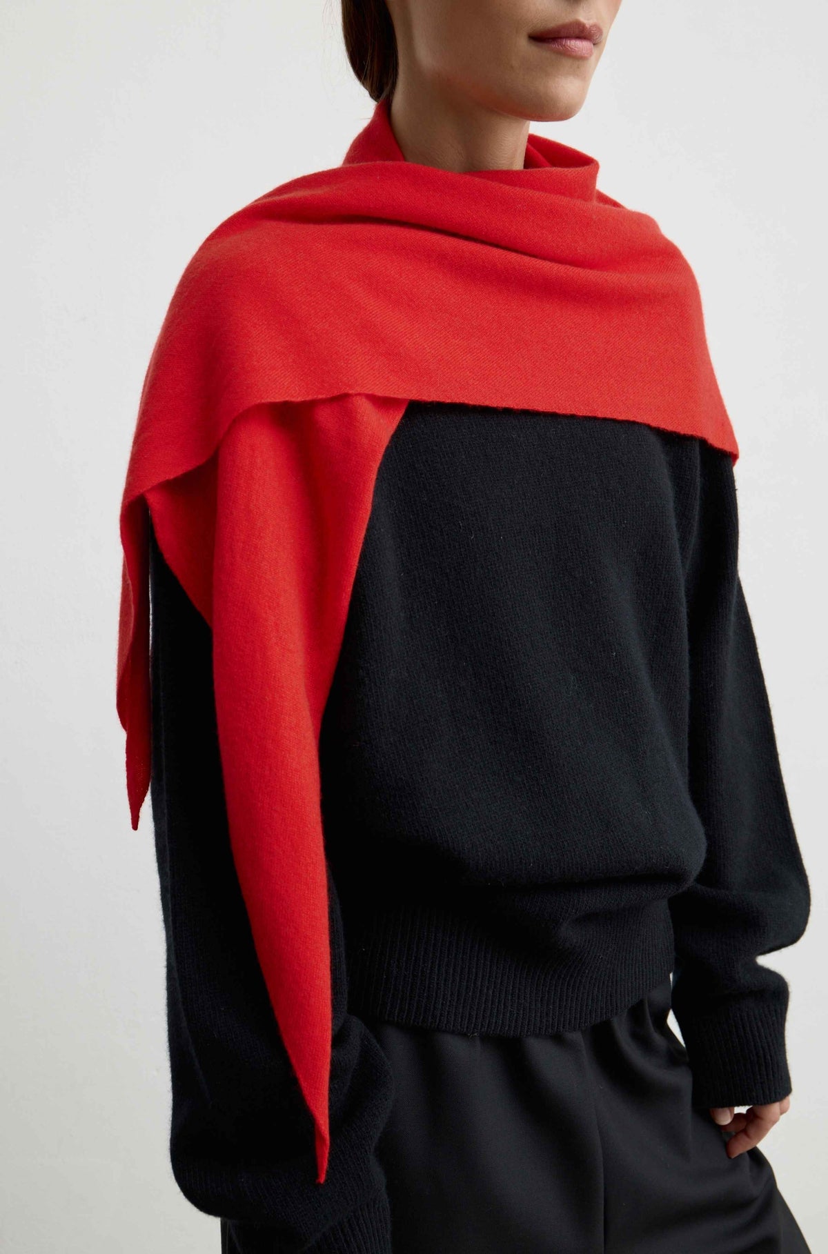 A close-up of a model wearing a vibrant red knit scarf wrapped around a black knit sweater against a plain light gray background.