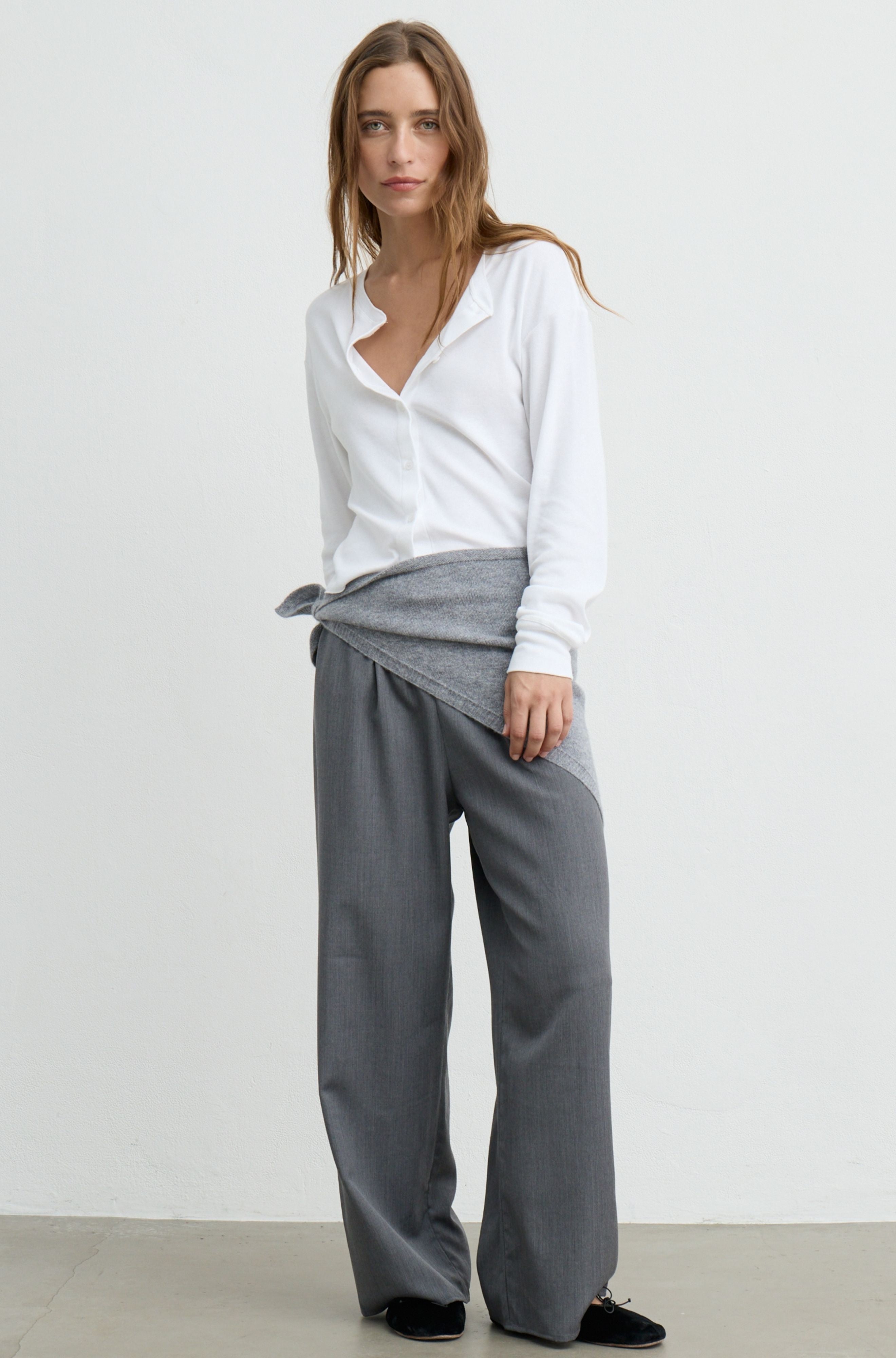 A woman modeling a white button-down shirt and wide-leg gray trousers, with a gray knit wrap tied around her waist, set against a plain white studio background.