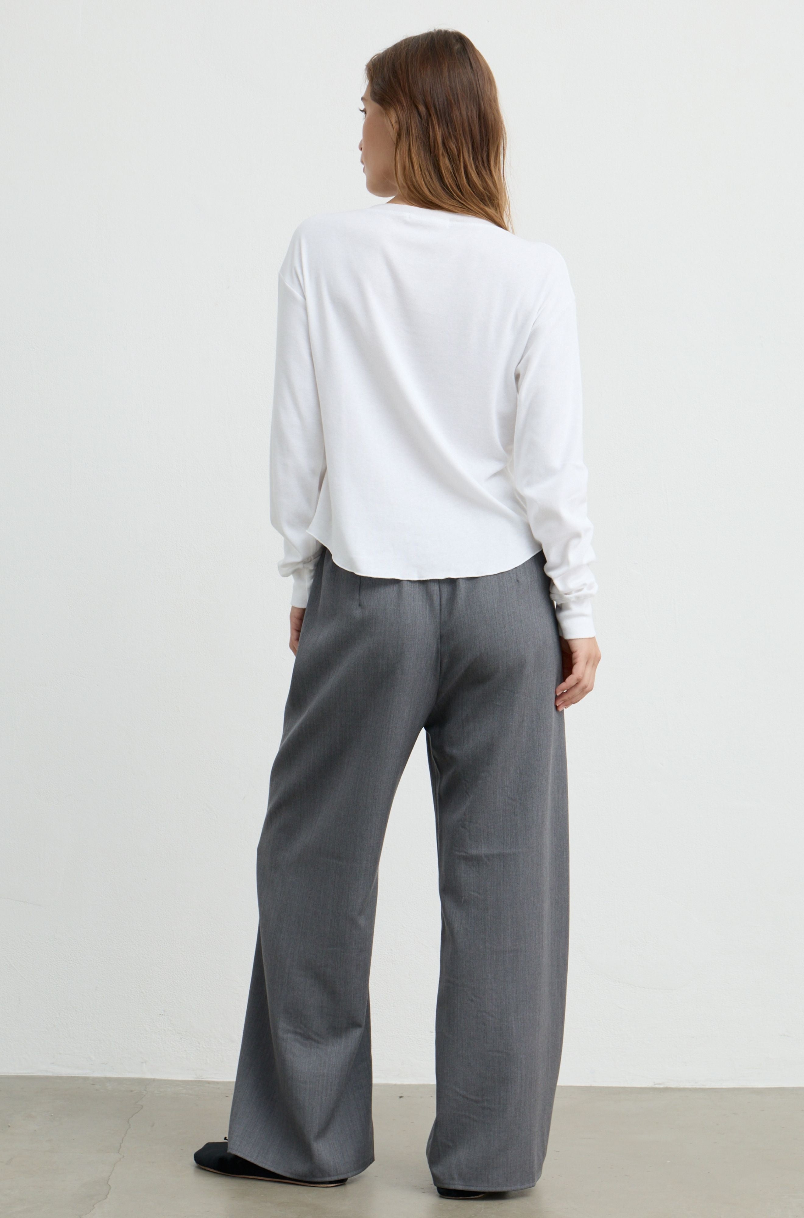 A woman shown from the back wearing a white long-sleeve top and gray wide-leg trousers, standing against a plain light gray wall.