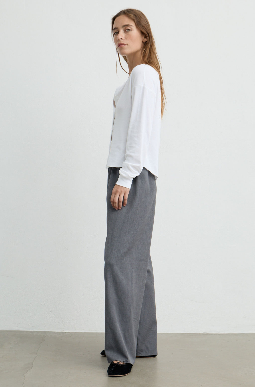 A woman stands in profile wearing a white long-sleeve top and wide-leg gray trousers against a plain light gray studio background.