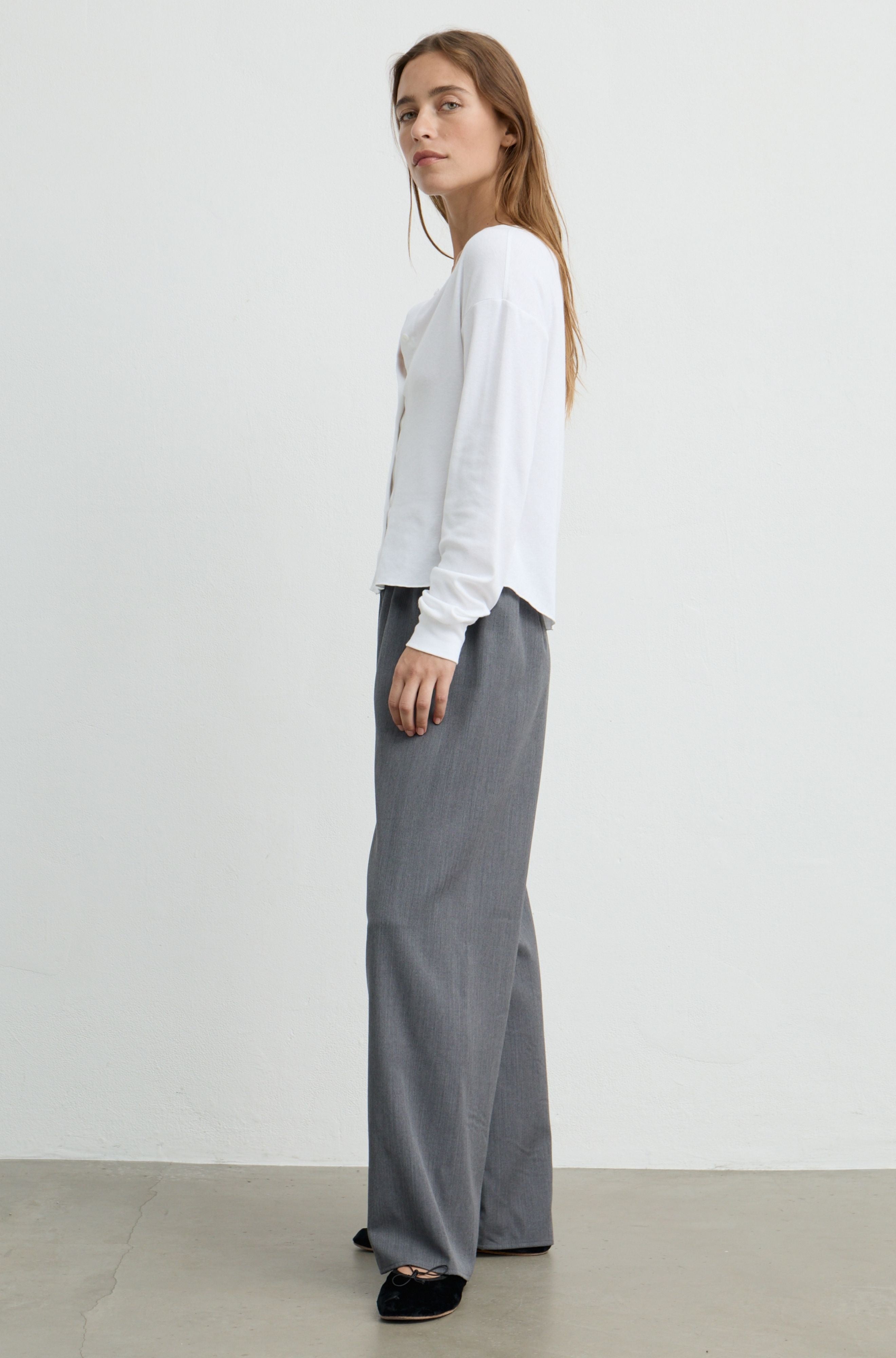 A woman stands in profile wearing a white long-sleeve top and wide-leg gray trousers against a plain light gray studio background.