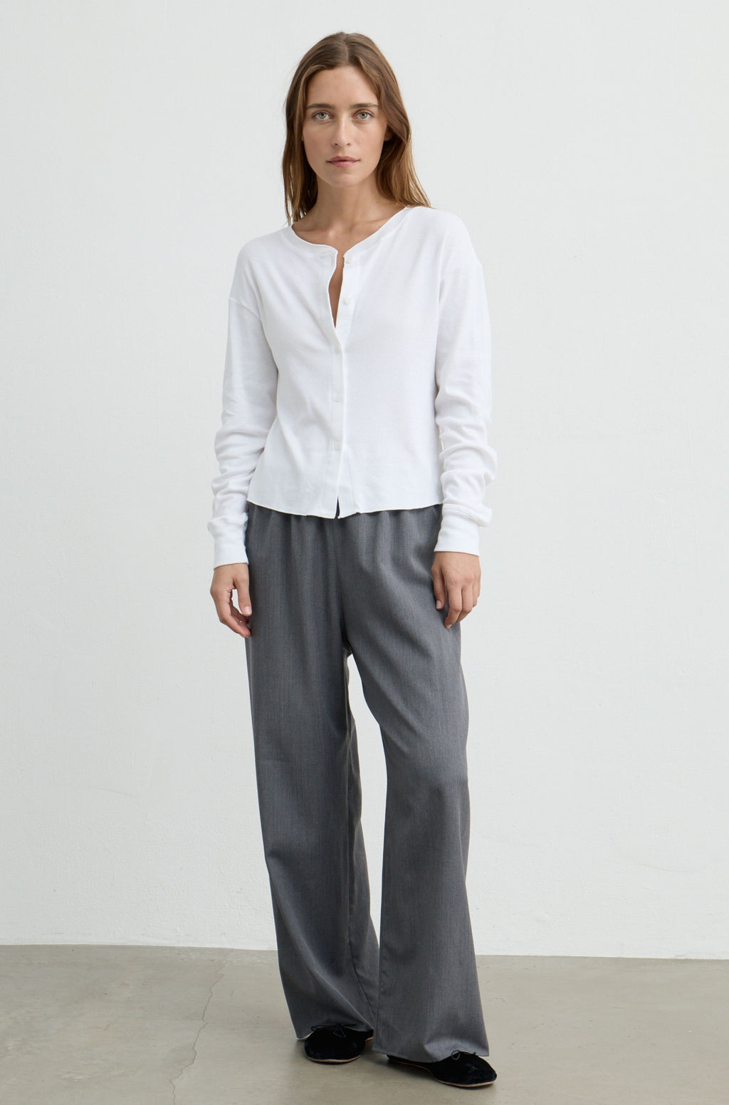 A woman modeling a white long-sleeve button-up shirt with a split neckline and grey wide-leg trousers, standing against a plain light gray studio background.