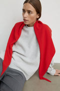 A woman wearing a bright red shawl draped over a light gray knit sweater and gray trousers, seated against a plain light gray wall.