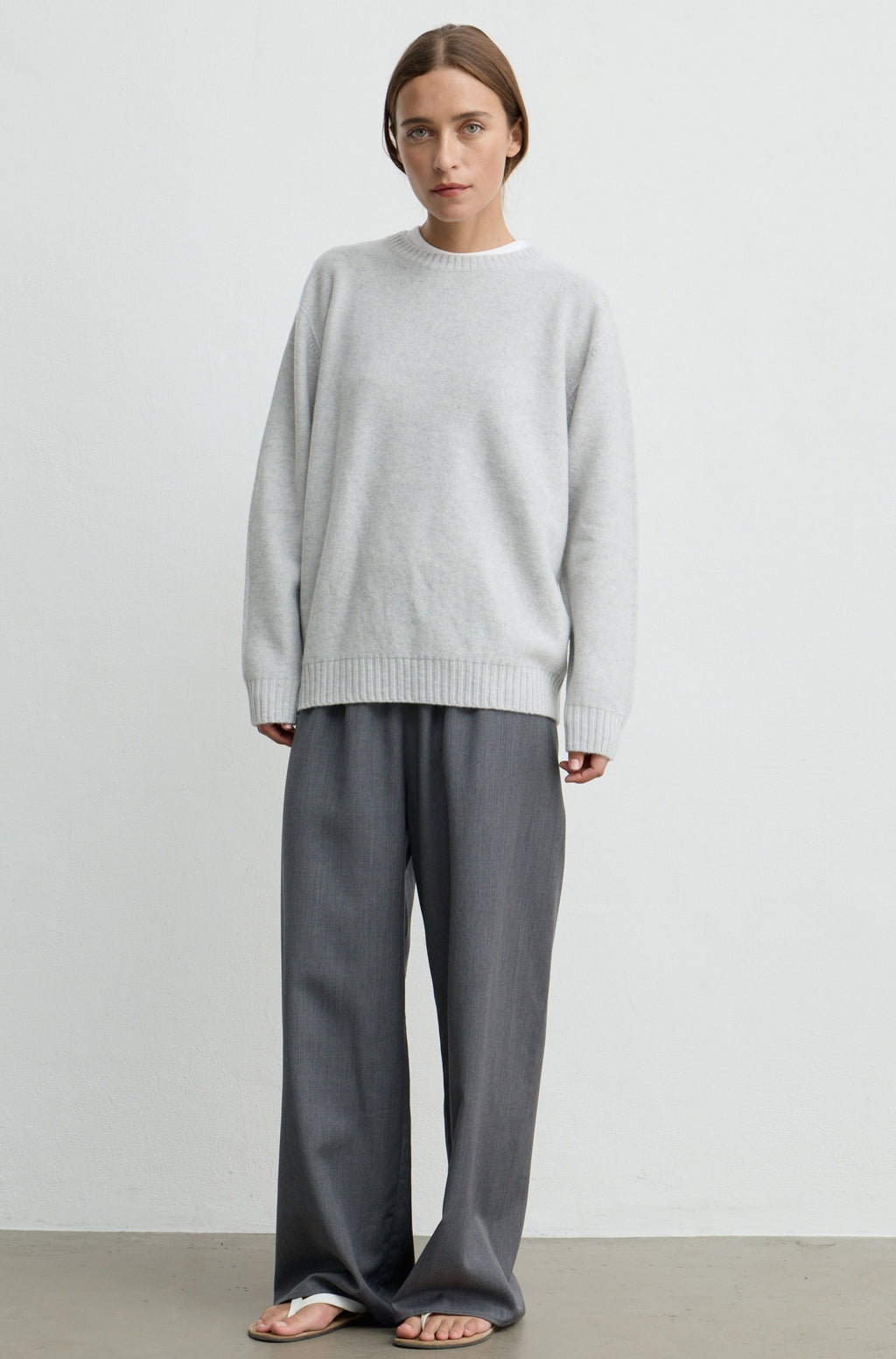 A woman stands against a plain light gray wall wearing an oversized light gray knit sweater, loose dark gray wide-leg pants, and sandals.