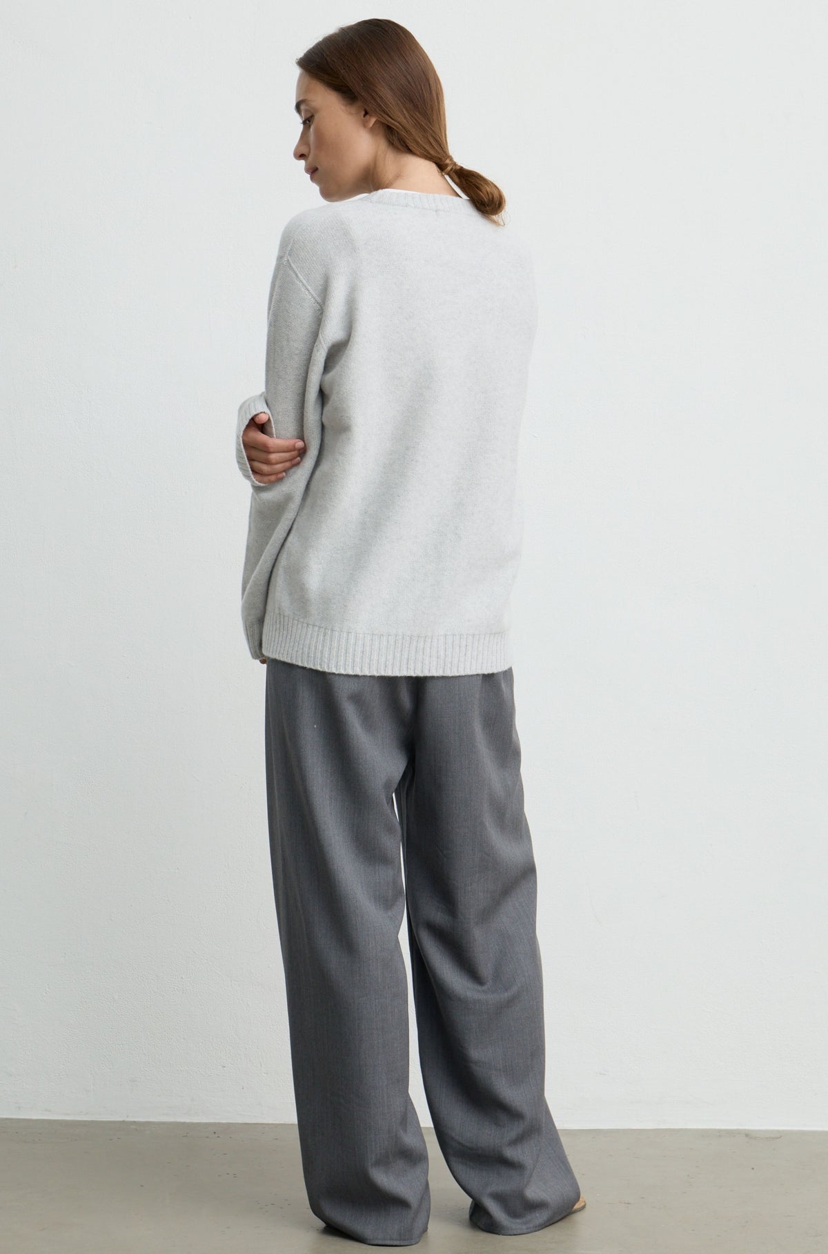 A woman shown from the back wearing a light gray knit sweater and wide-leg gray trousers, standing against a plain white wall.