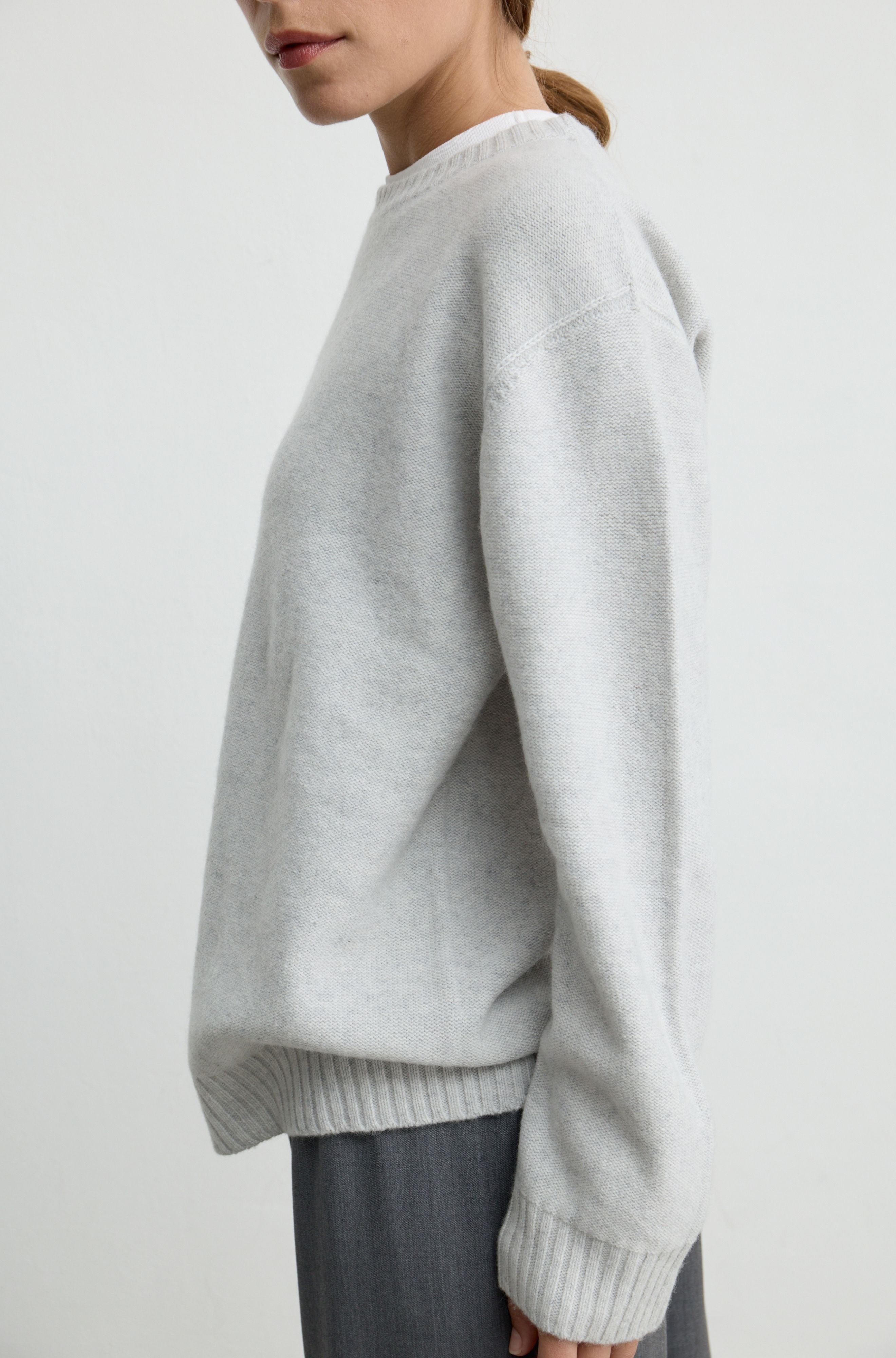 A close-up side view of a person wearing a light gray knit sweater with a crew neck and ribbed hem and cuffs, paired with dark gray pants, against a plain light gray background.