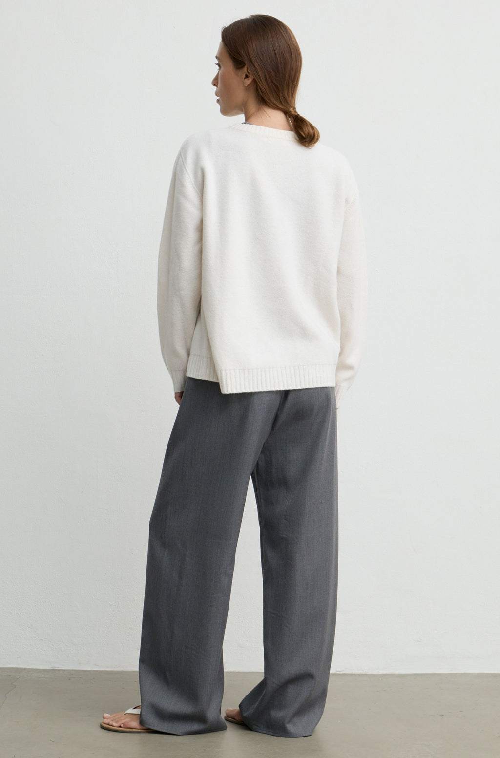 A woman shown from the back wearing a cream knit sweater and wide-leg gray trousers, standing against a plain light gray wall.