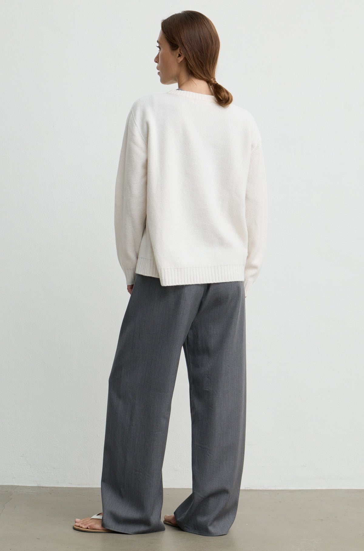 A woman shown from the back wearing a cream knit sweater and wide-leg gray trousers, standing against a plain light gray wall.