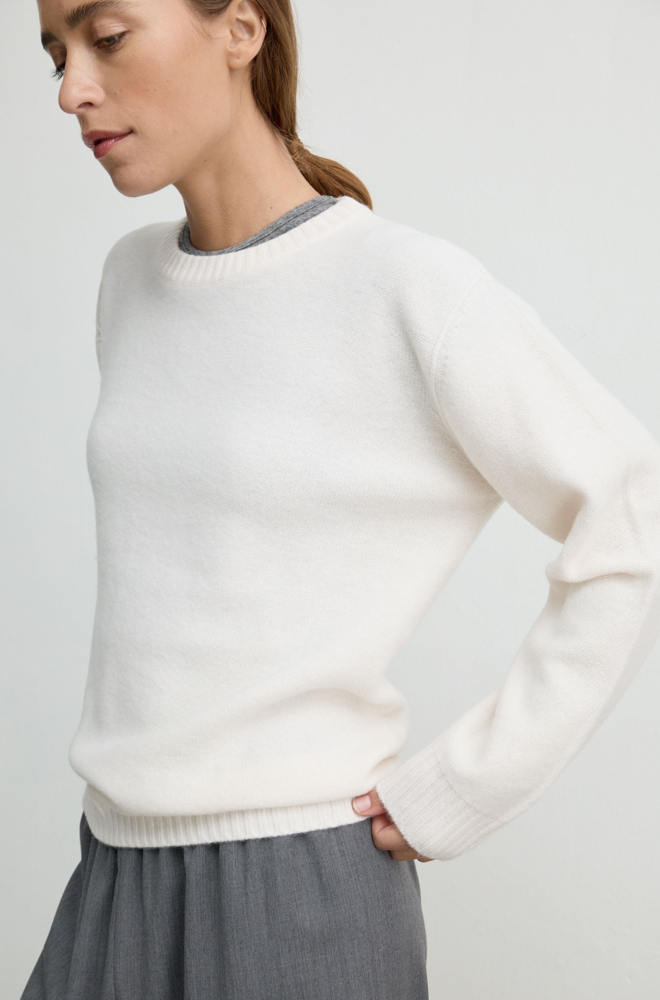 A woman wearing a cream-colored knit crewneck sweater with a gray ribbed inner collar, paired with a gray skirt, against a light gray studio background.